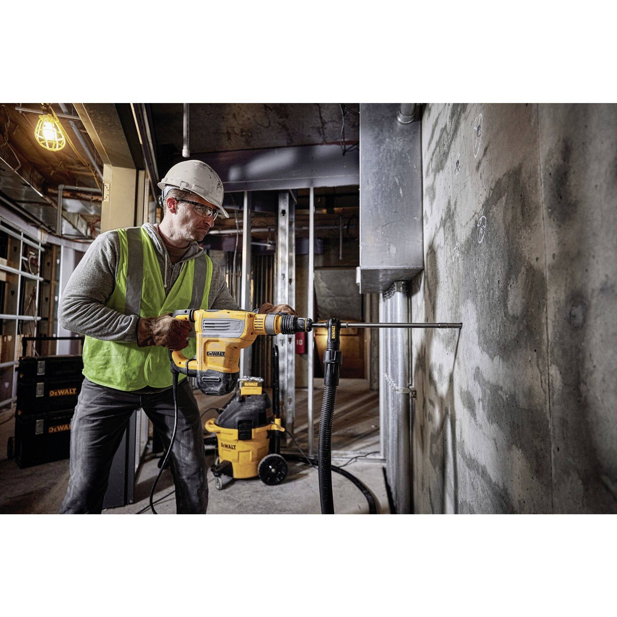 Un ouvrier en tenue de sécurité fore un mur en béton sur un chantier, entouré d'échafaudages et d'outils.