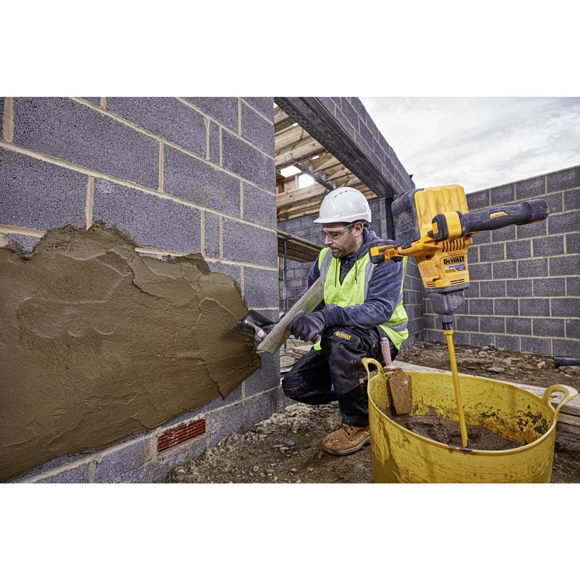 Un ouvrier enduit un mur de briques avec une truelle. À côté de lui se trouve un mélangeur de mortier jaune. Des vêtements de sécurité sont portés.