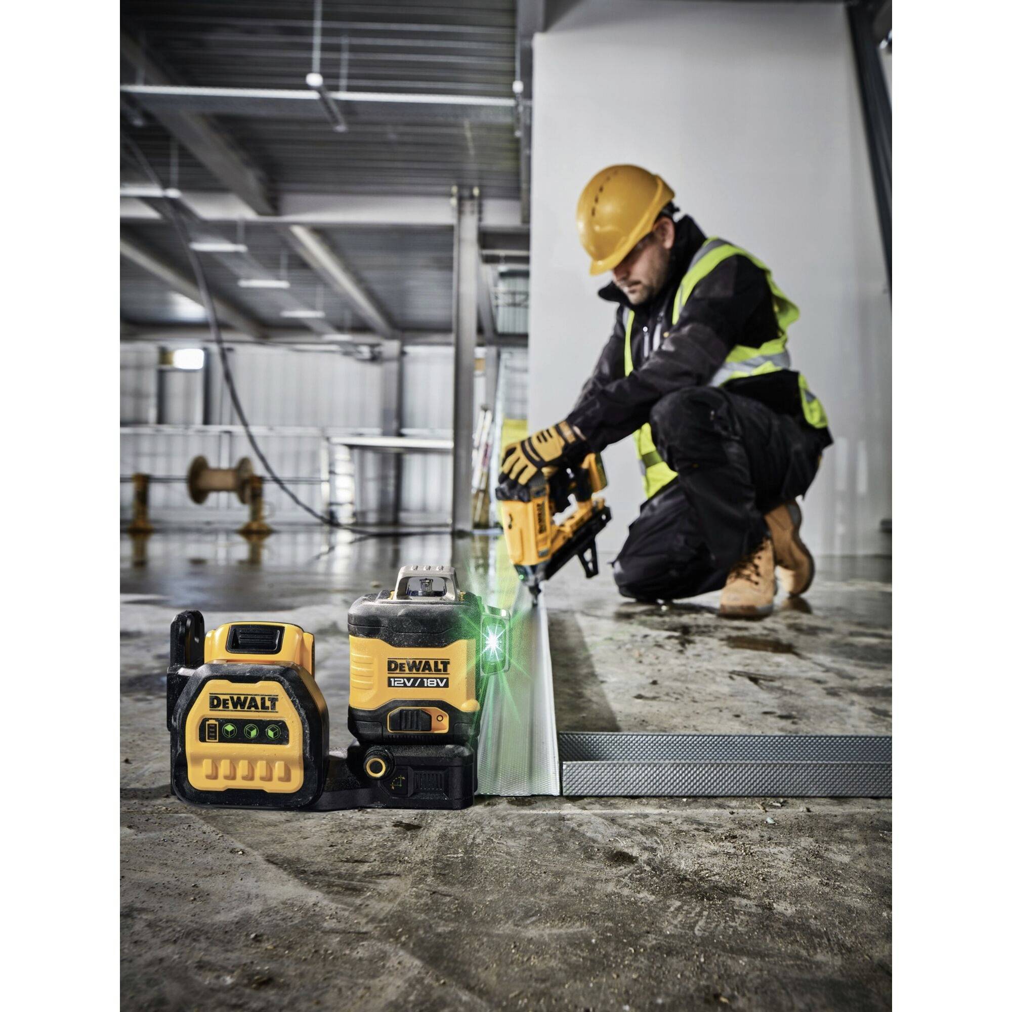 Un ouvrier portant un casque de protection et un gilet de sécurité utilise un cloueur sans fil sur un chantier de construction. Des outils Dewalt sont visibles au premier plan.