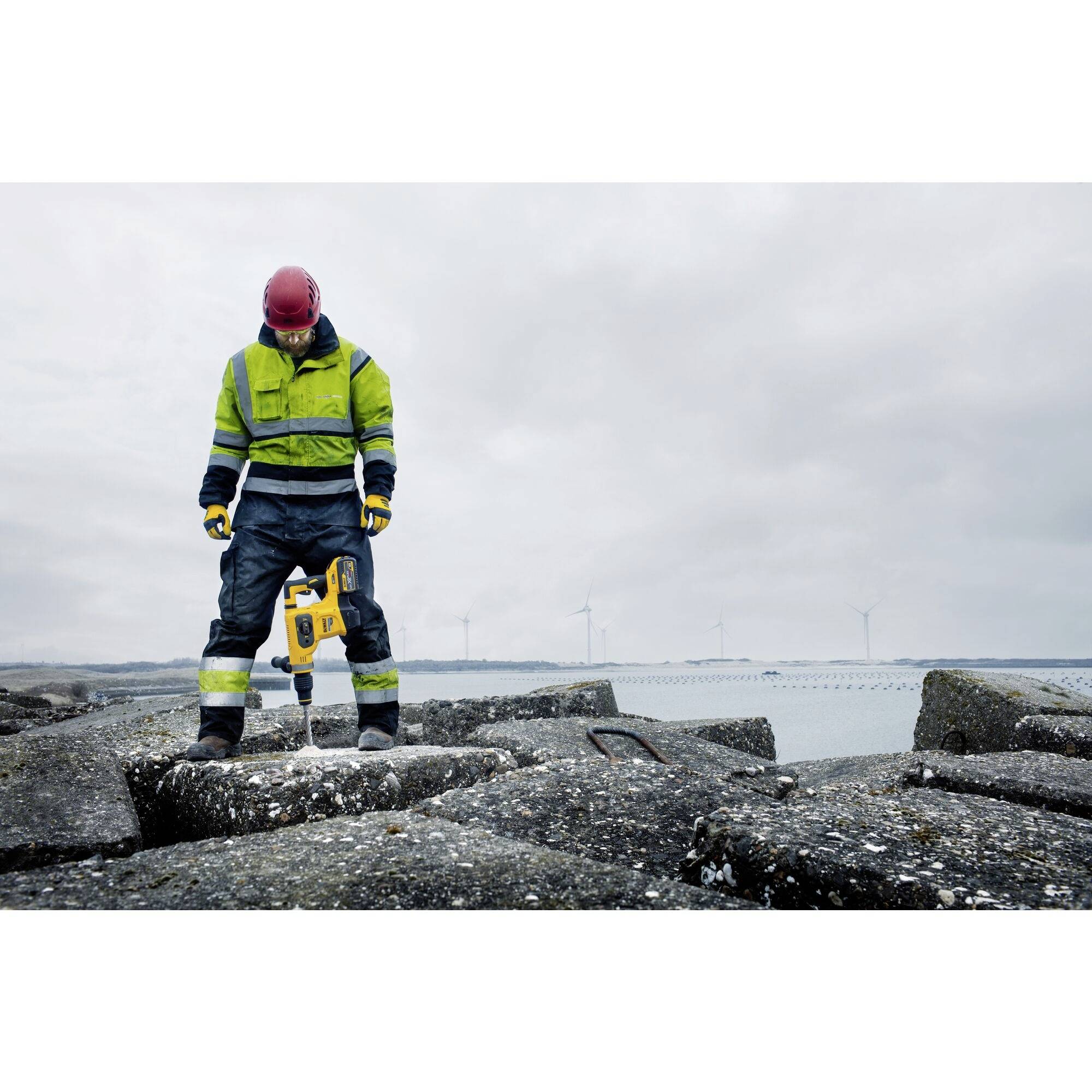 Un ouvrier du bâtiment portant un gilet de sécurité et un casque travaille avec un marteau-piqueur sur de grands blocs de béton près de la côte, avec des éoliennes en arrière-plan.
