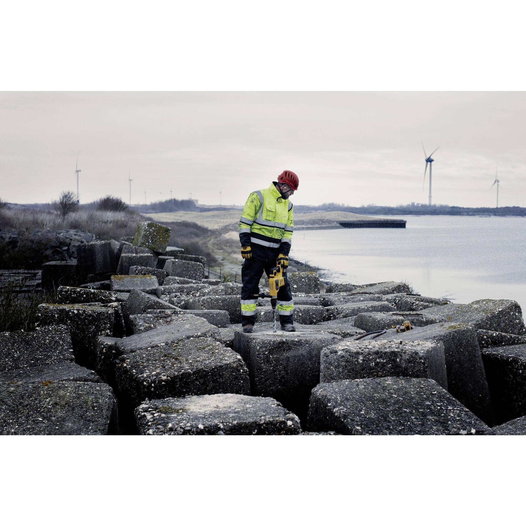 Une personne en combinaison de protection avec des bottes debout sur de grands blocs de béton près d'un plan d'eau, des éoliennes et un ciel nuageux en arrière-plan.