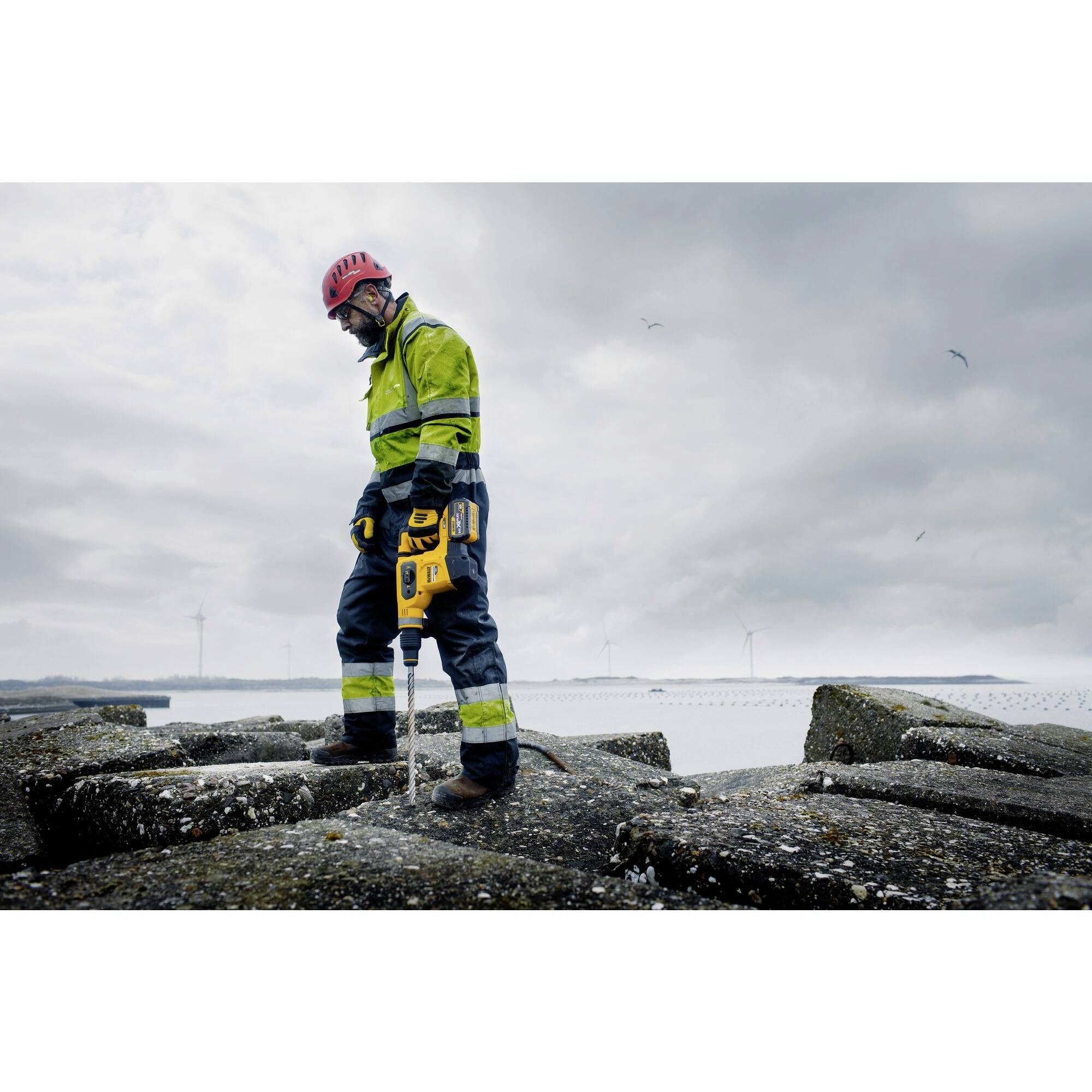 Un travailleur portant un équipement de protection et un casque se tient sur un terrain rocailleux et fore avec un appareil jaune. Des éoliennes sont visibles en arrière-plan.