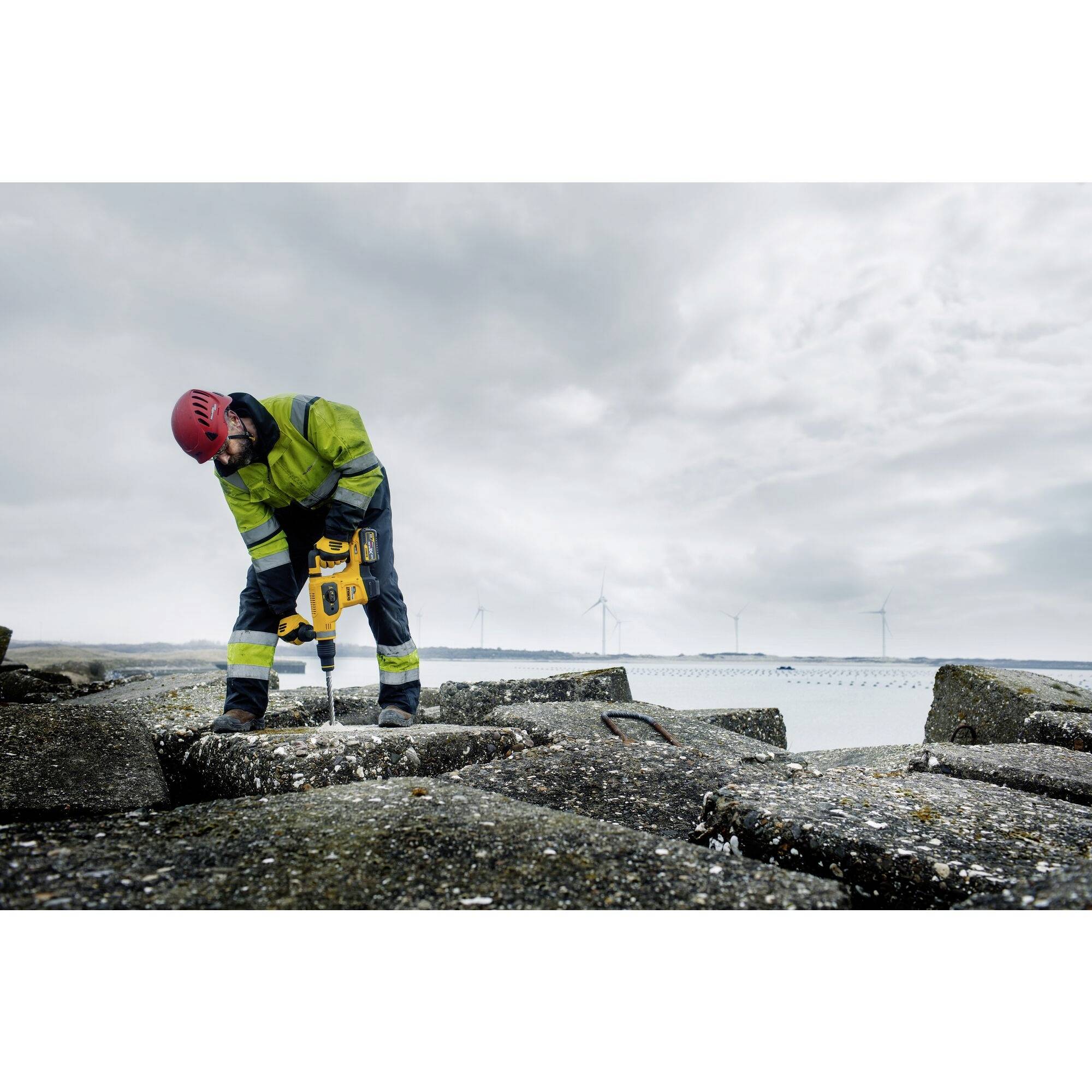 Un ouvrier portant un casque rouge et des vêtements de sécurité fore une surface en béton à l'aide d'une machine. Des éoliennes sont visibles en arrière-plan.