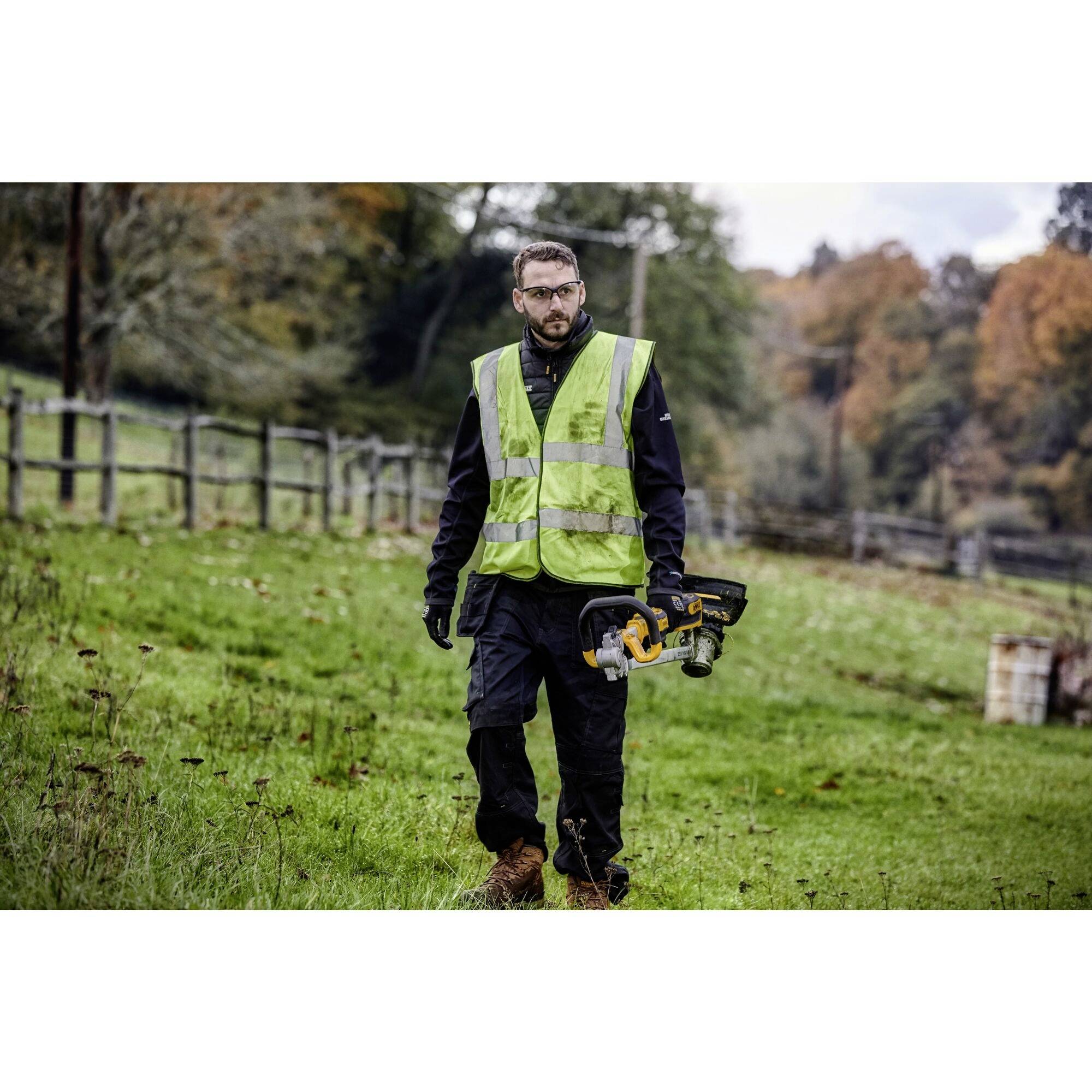 Une personne dans un champ porte un gilet de sécurité et tient un outil électrique à la main. L'arrière-plan montre des arbres en automne.