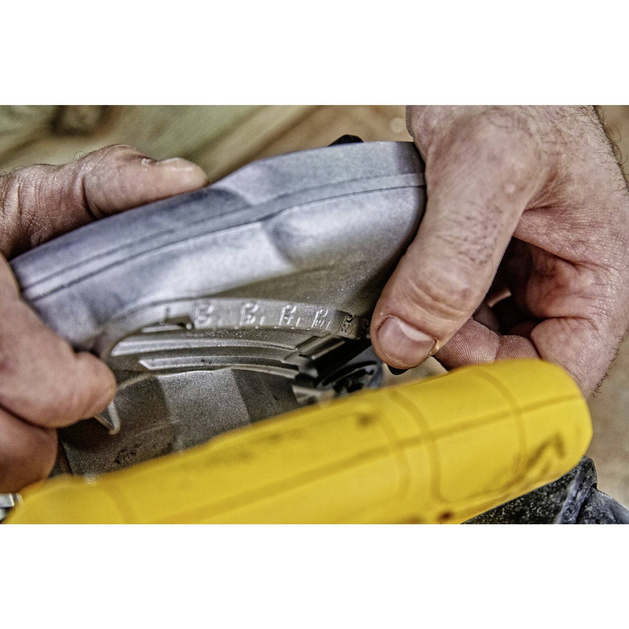 Deux mains tiennent une pièce métallique avec une machine jaune. Des travaux ou des réparations pourraient être effectués sur un appareil.