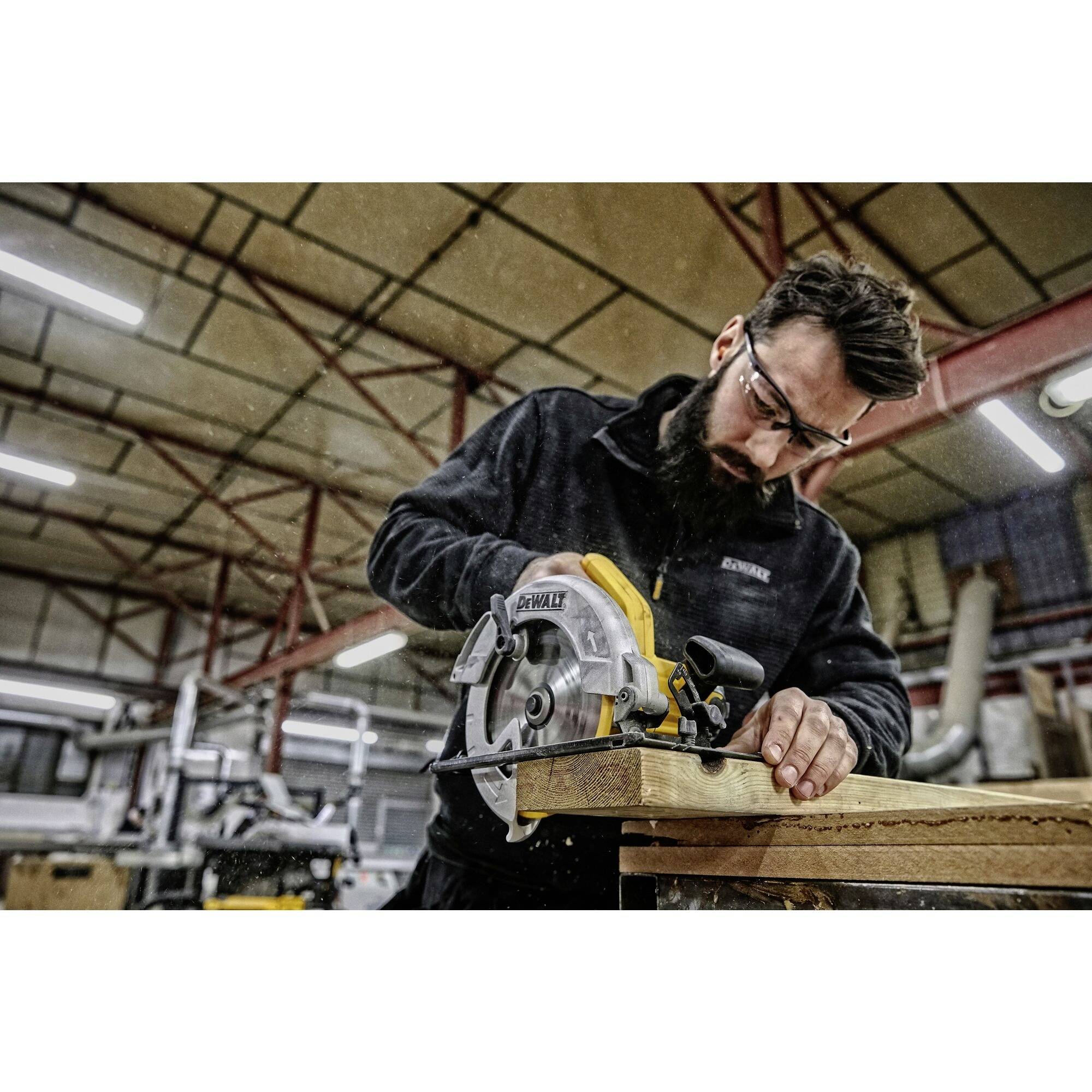 Un homme portant des lunettes de protection coupe du bois avec une scie circulaire dans un atelier. Des machines et des étagères sont visibles en arrière-plan.