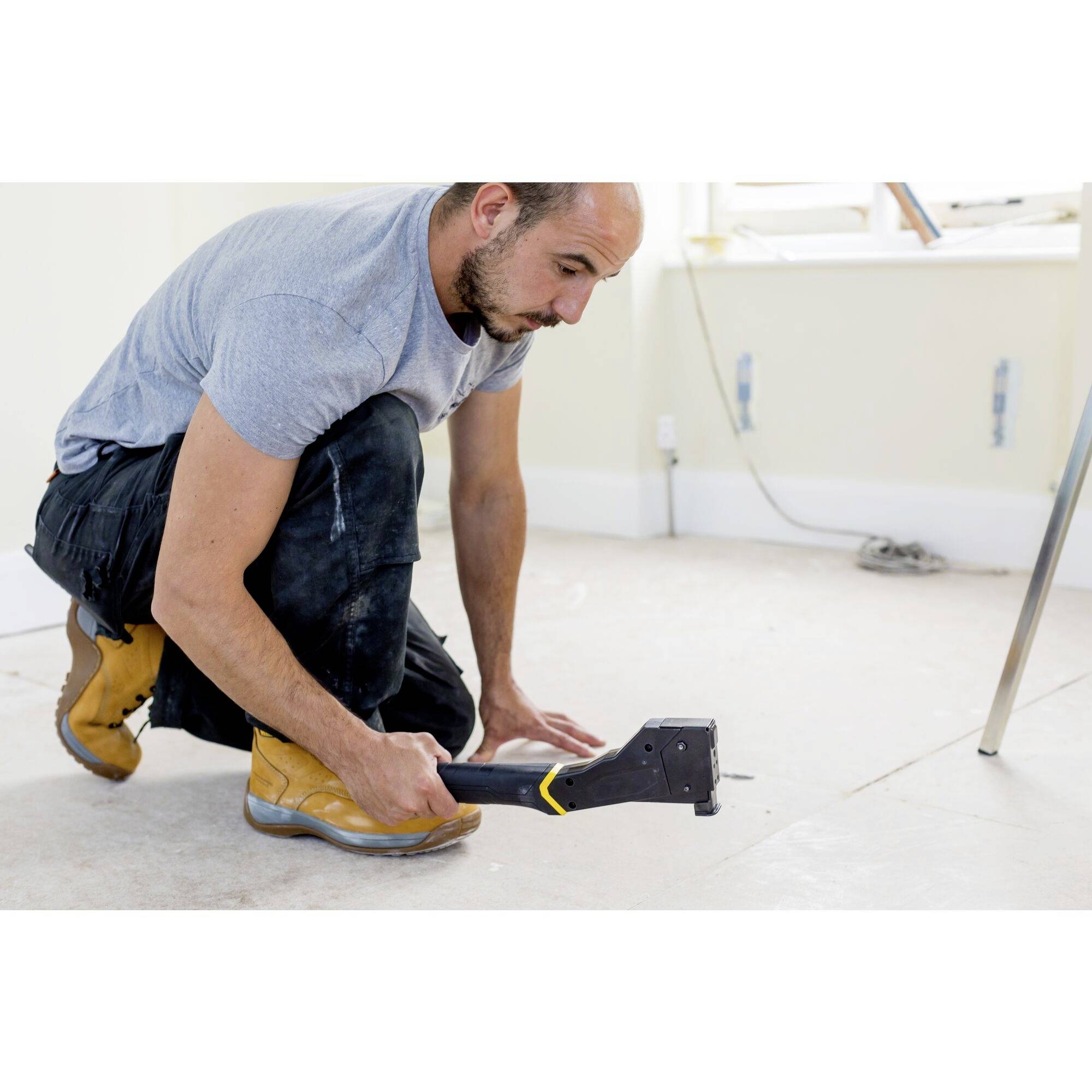Un homme est agenouillé sur un sol et utilise un agrafeur manuel pour fixer un tapis. Il porte des vêtements de travail et des bottes.