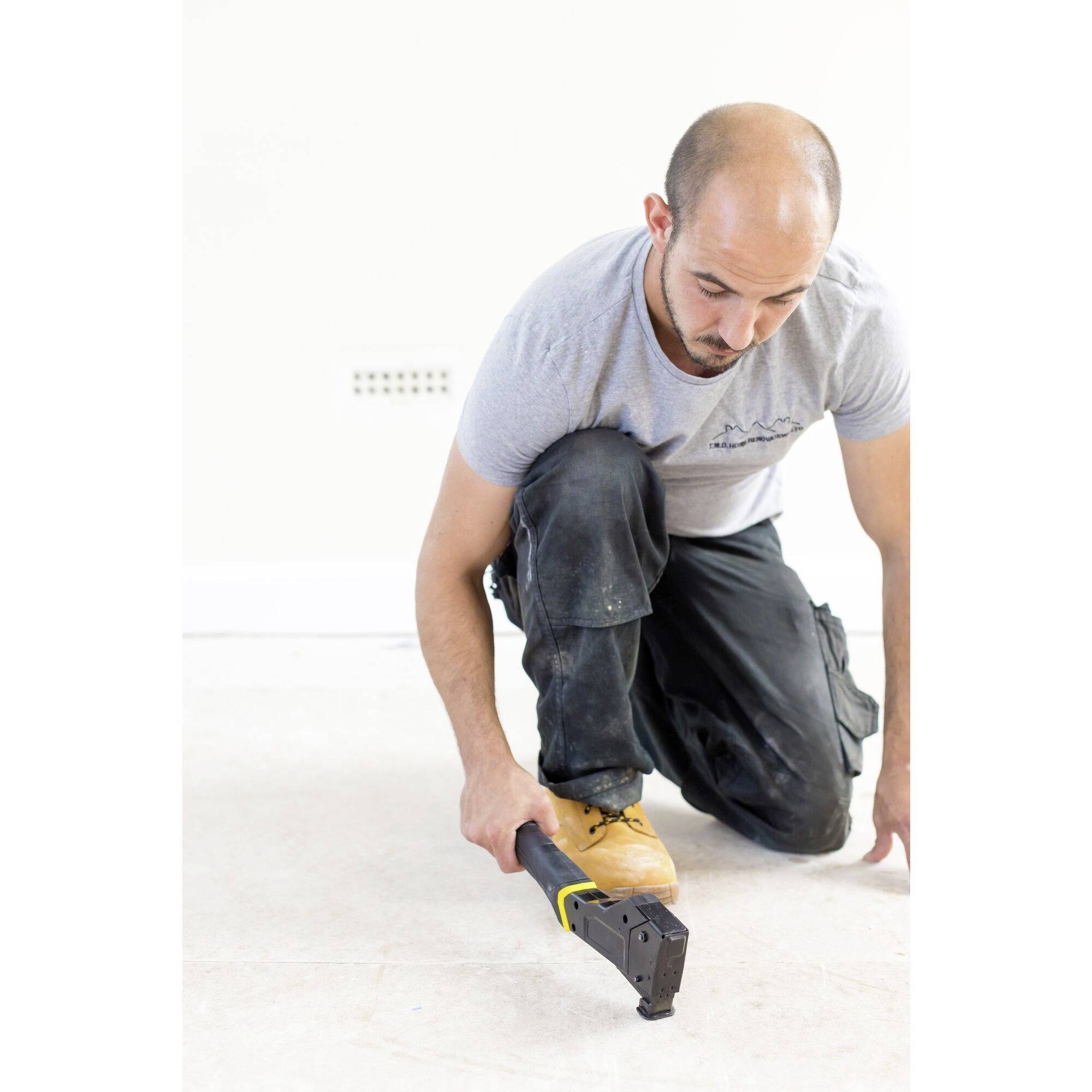 Un homme est agenouillé par terre et travaille avec un outil manuel qui a un manche jaune. Il porte un t-shirt gris et un pantalon foncé.