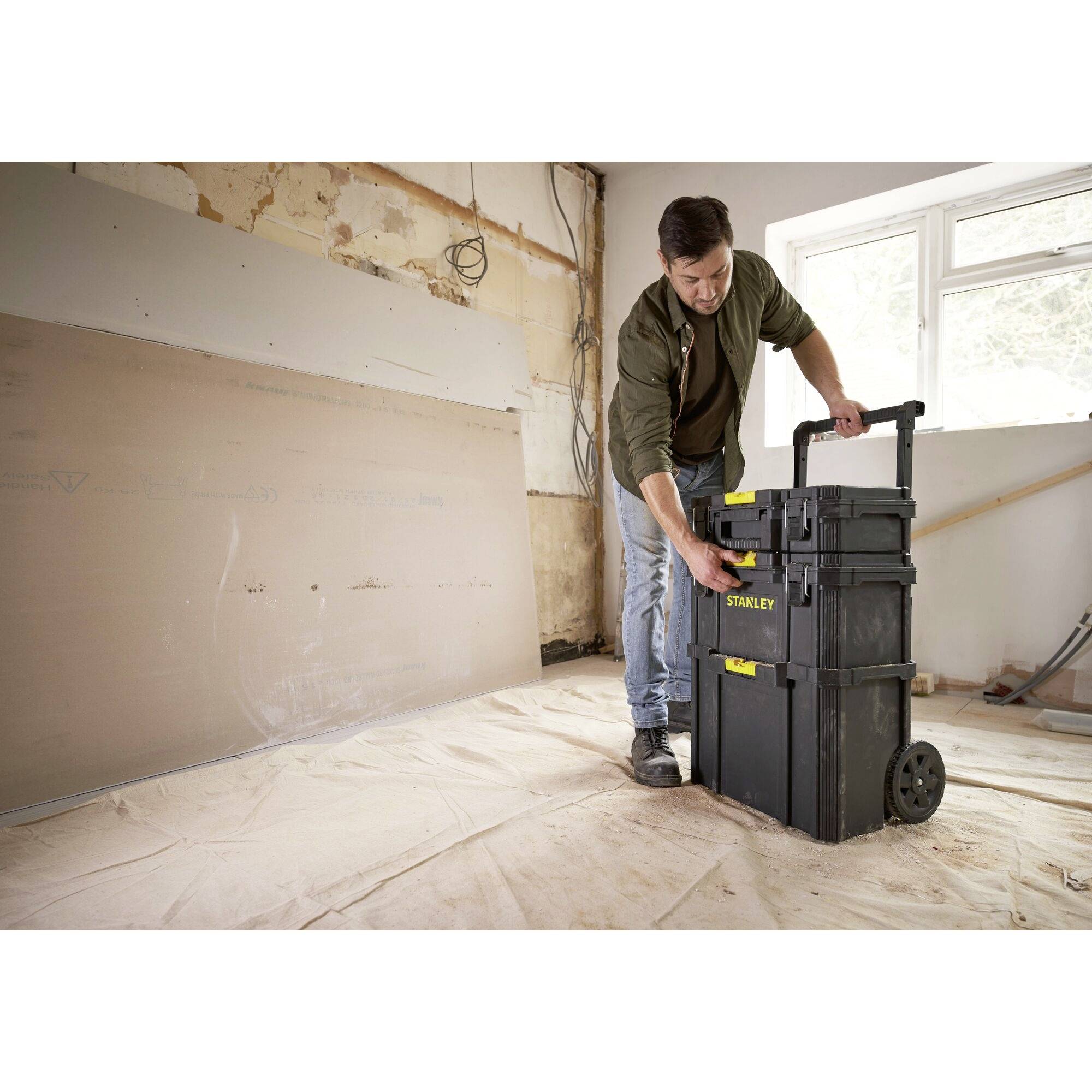 Un homme se tient dans une pièce partiellement rénovée et soulève une boîte à outils noire sur roulettes. Des plaques de plâtre sont visibles en arrière-plan.