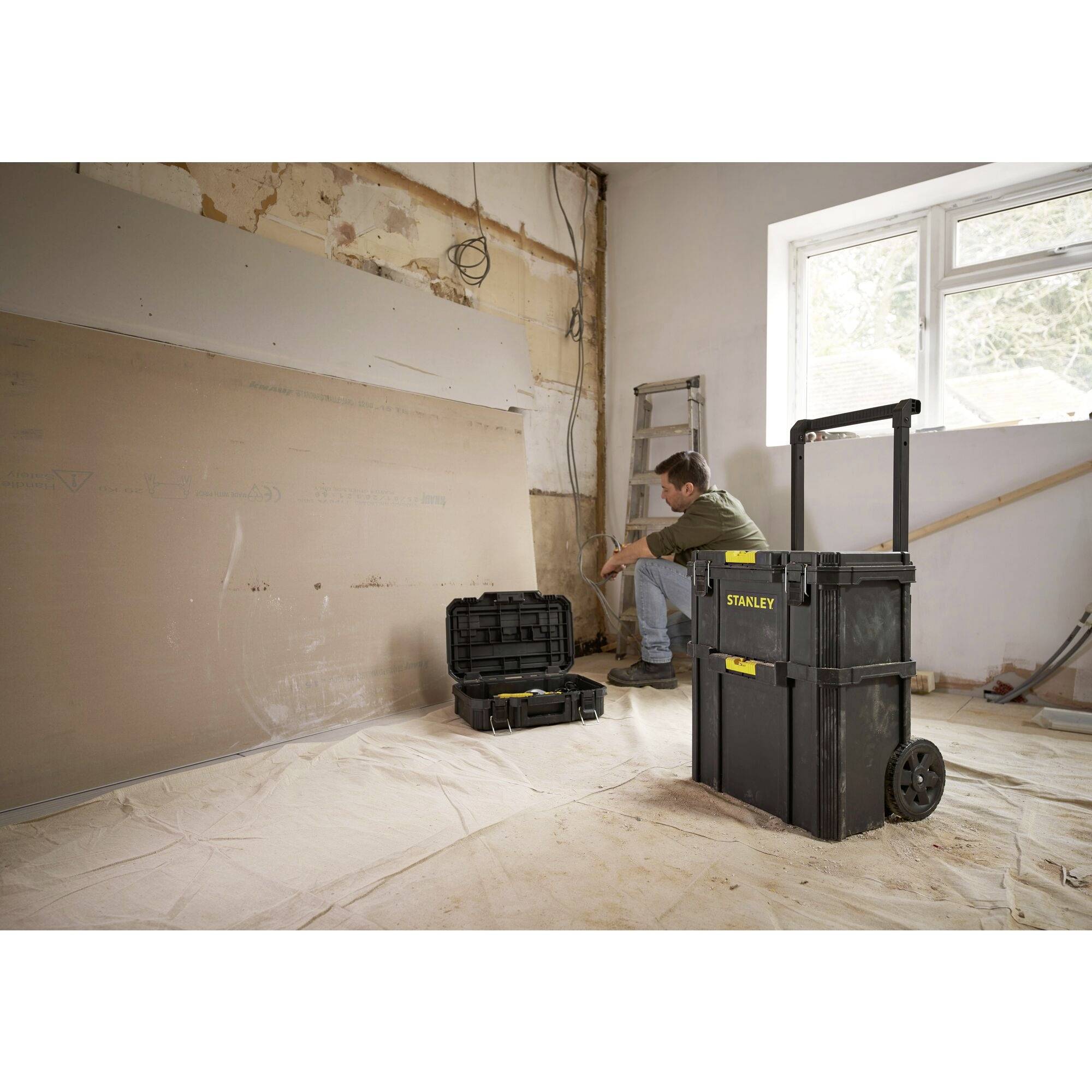 Un homme dans une pièce avec une boîte à outils et une échelle, travaille sur un chantier. Les murs sont partiellement démontés, le sol est recouvert d'une bâche.
