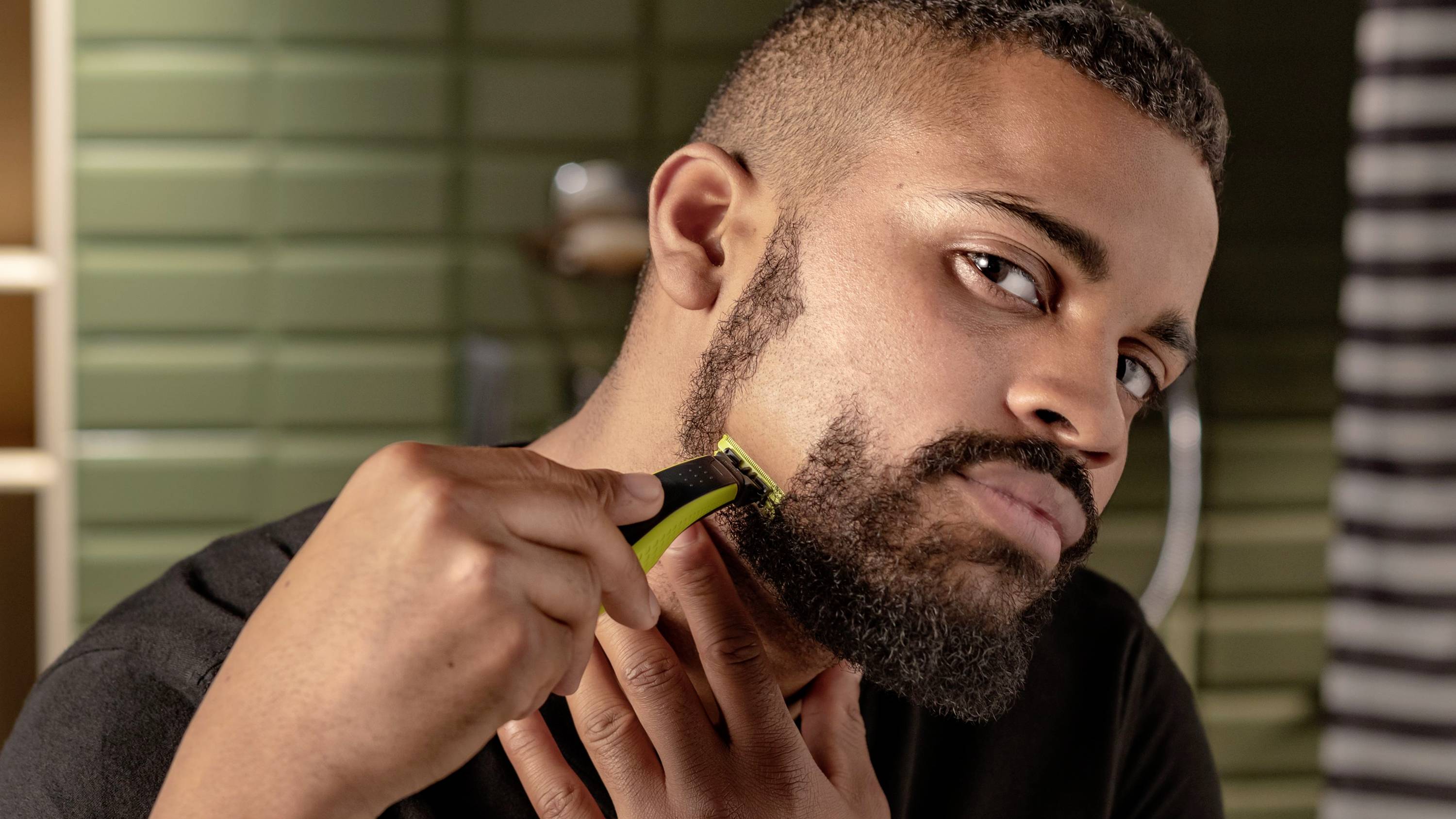 Un homme se rase la barbe avec un rasoir électrique dans une salle de bain. Des carreaux verts sont visibles en arrière-plan.