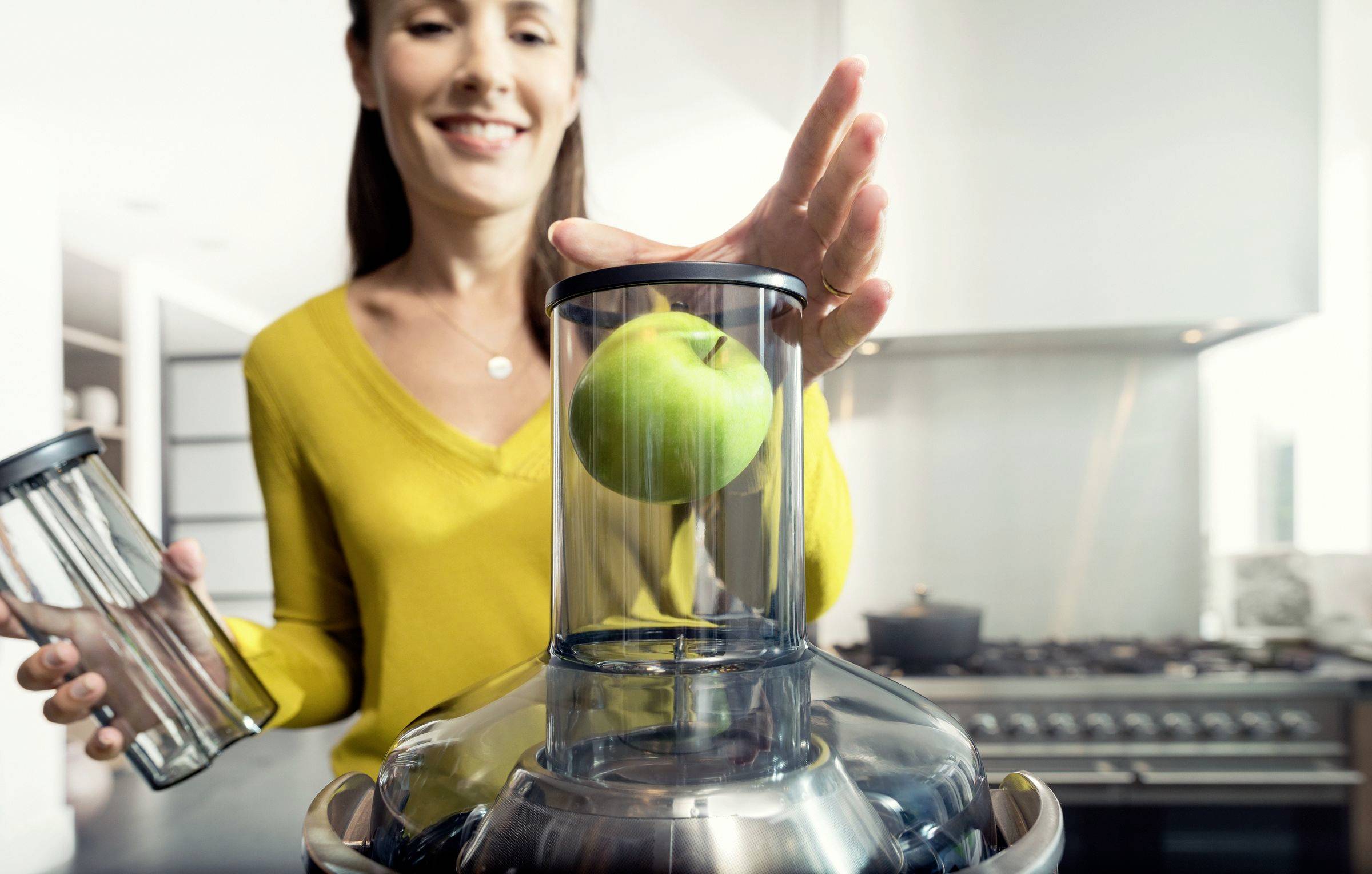 Une femme place une pomme verte dans un extracteur de jus dans une cuisine, prête à préparer du jus. Elle tient un récipient dans son autre main.