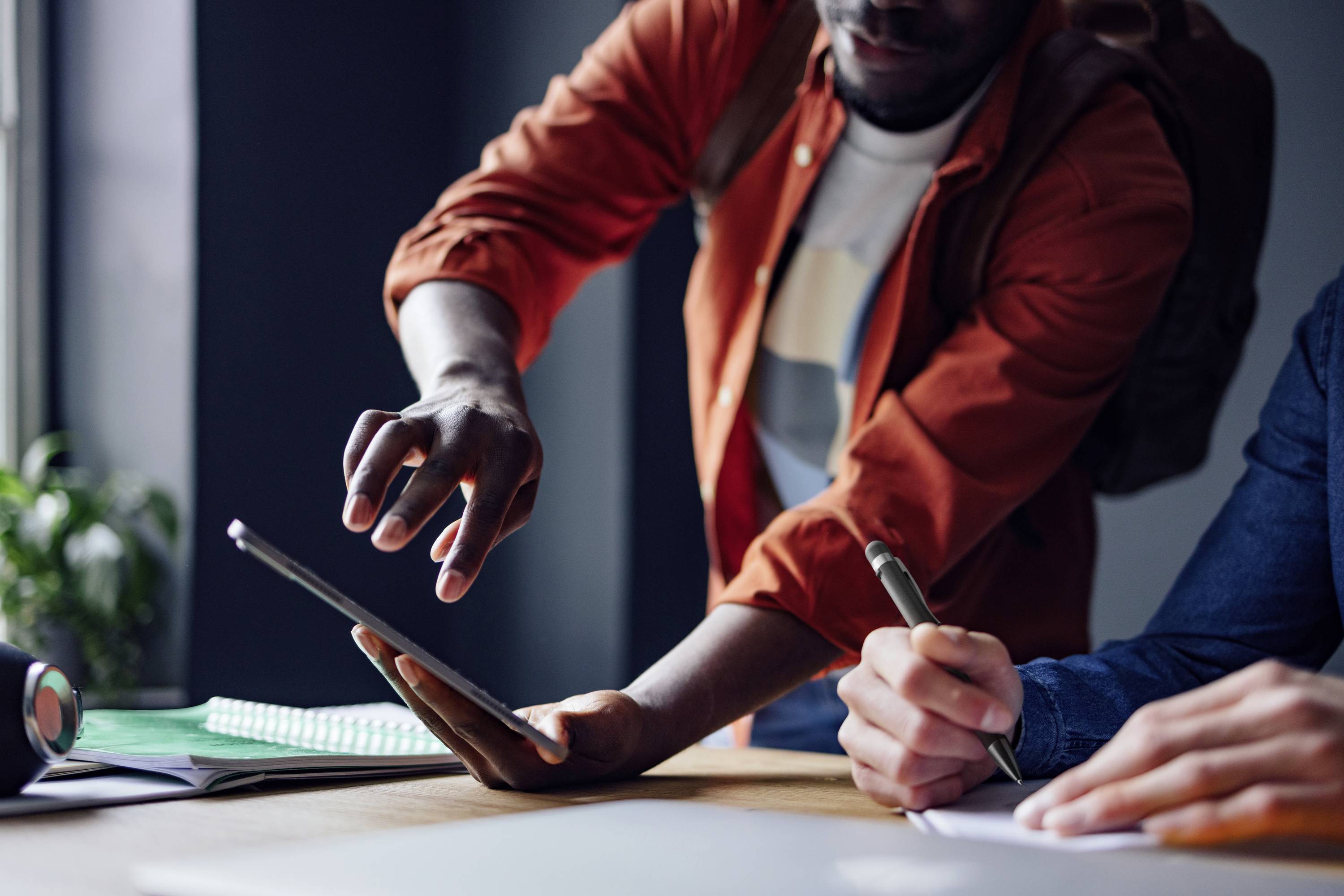 Deux personnes travaillent à une table, l'une montrant quelque chose sur une tablette tandis que l'autre prend des notes. Ambiance de bureau baignée de lumière solaire.