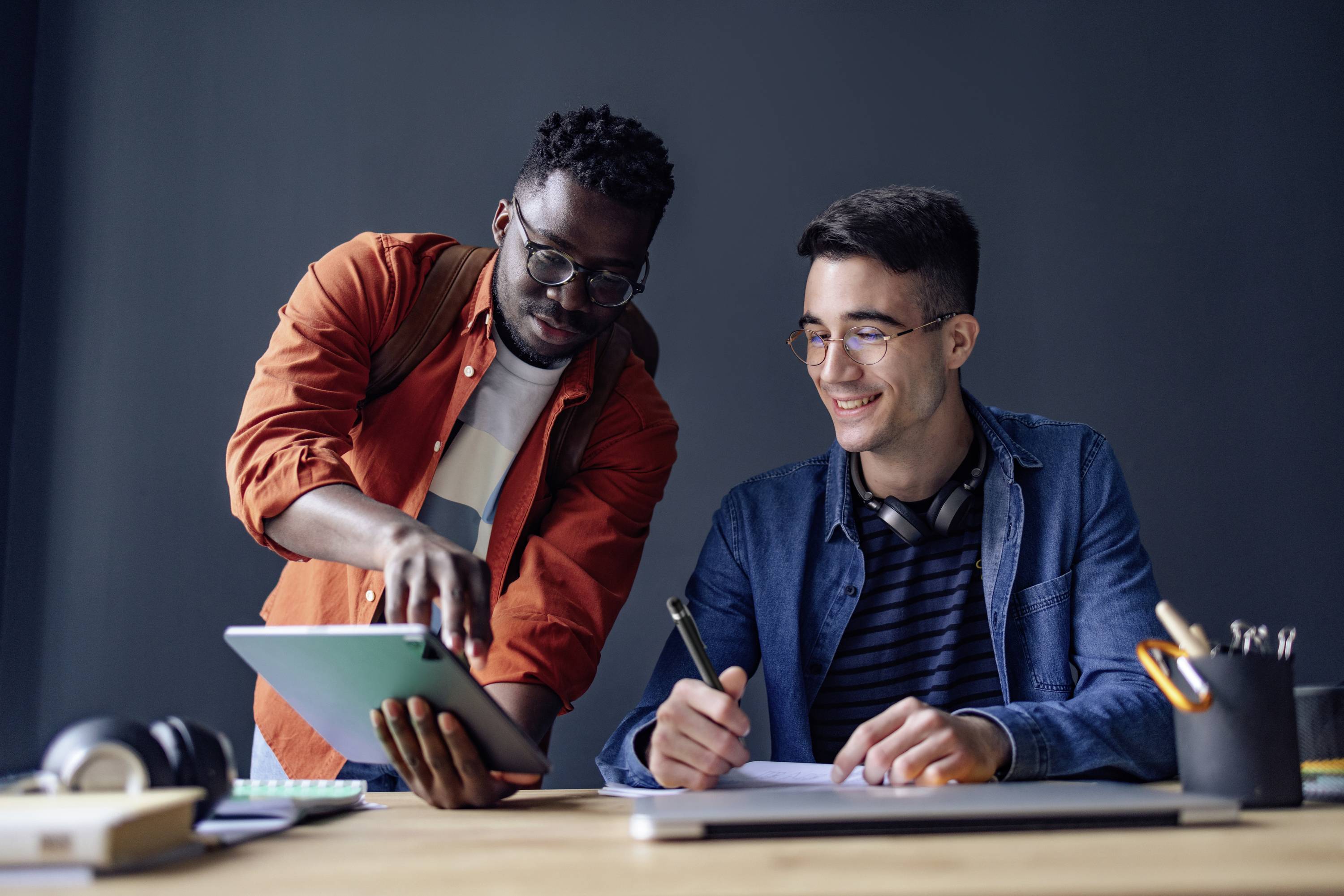 Deux hommes travaillent ensemble à une table ; l'un pointe un tablet, l'autre prend des notes.