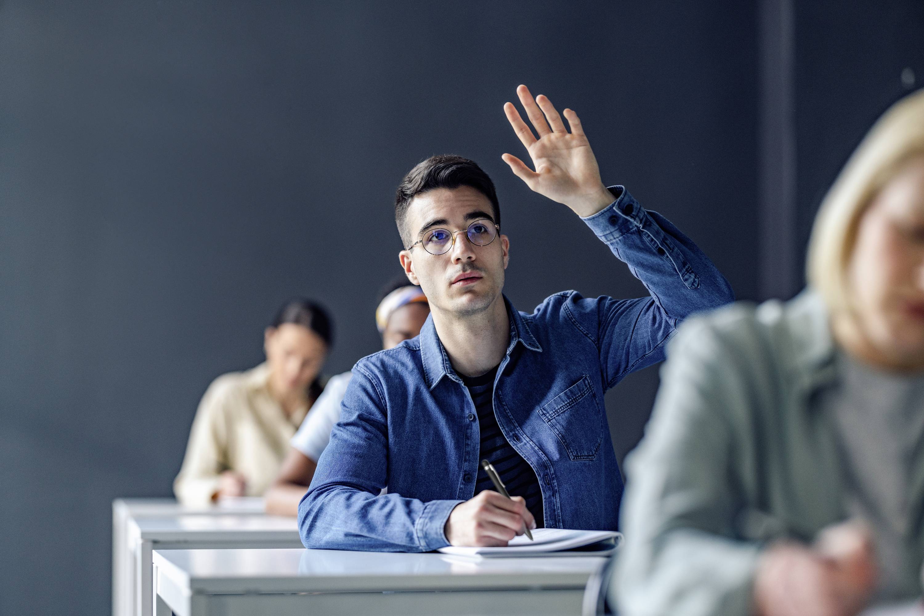 Un jeune homme lève la main alors qu'il est assis à un bureau dans une salle de classe. D'autres élèves sont flous en arrière-plan.