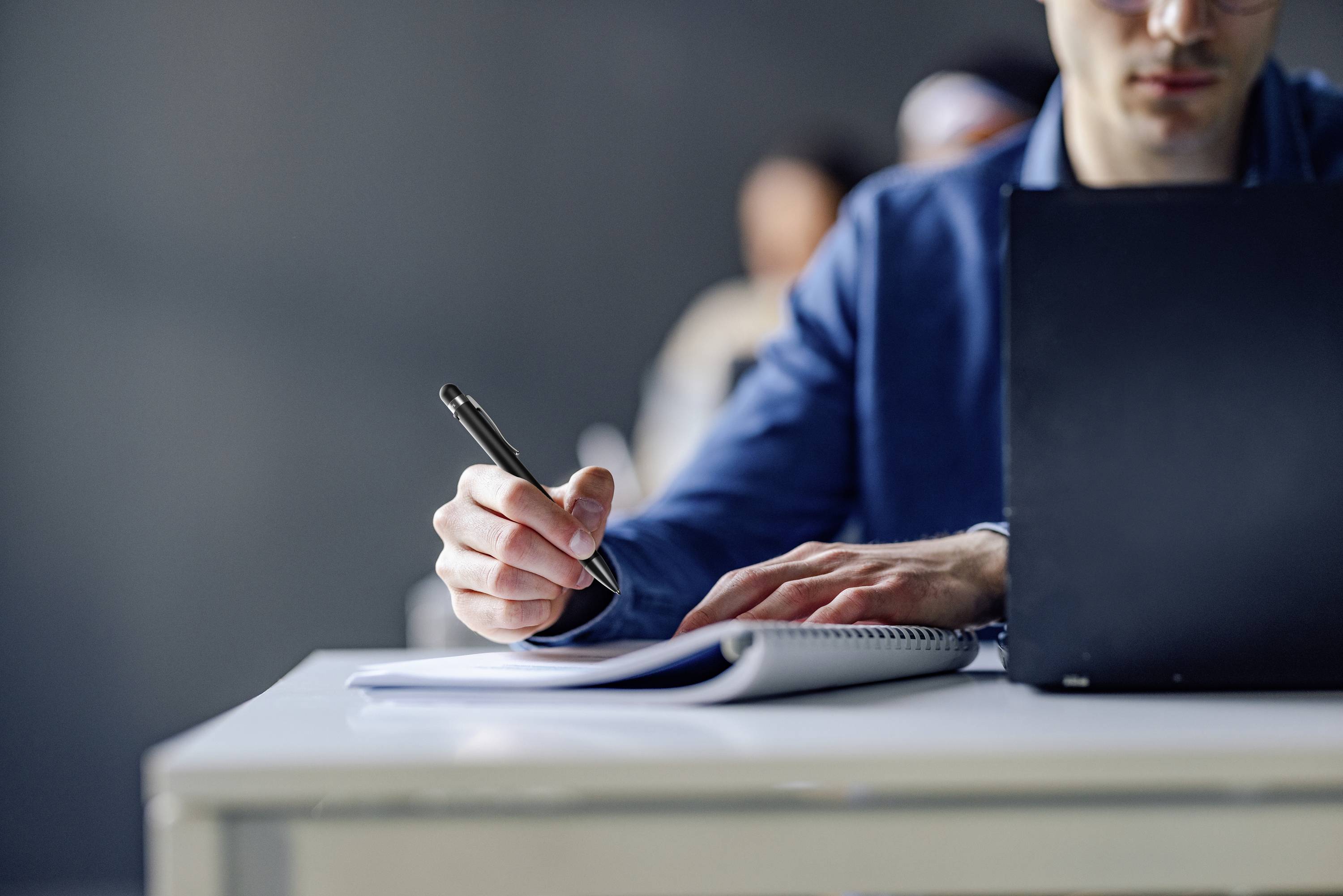 Une personne portant un costume bleu écrit dans un carnet à une table, à côté d'un ordinateur portable ouvert. L'arrière-plan est flou.