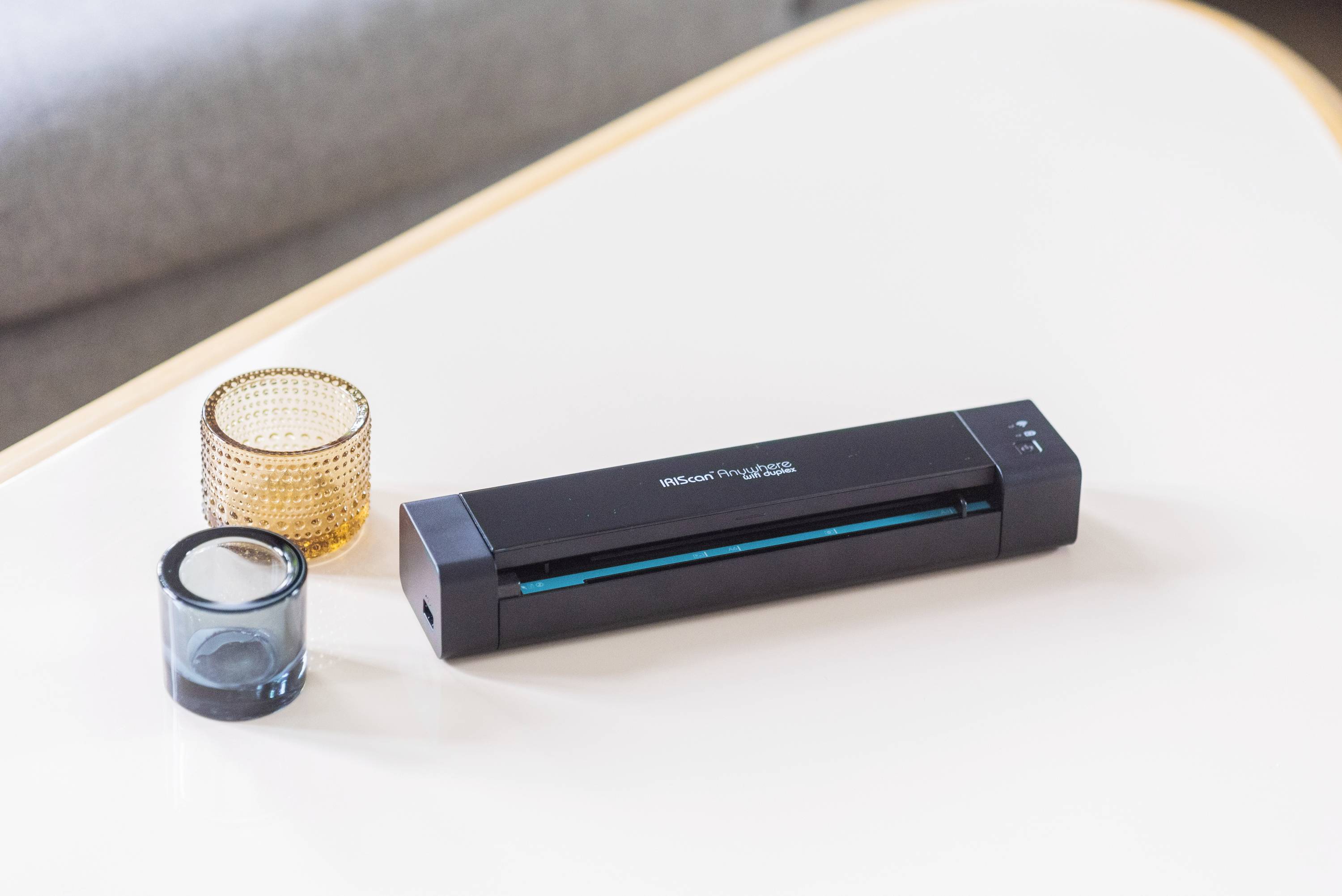 Un scanner portable est posé sur une table à côté de deux récipients décoratifs en verre. Il est compact et adapté à la numérisation de documents.
