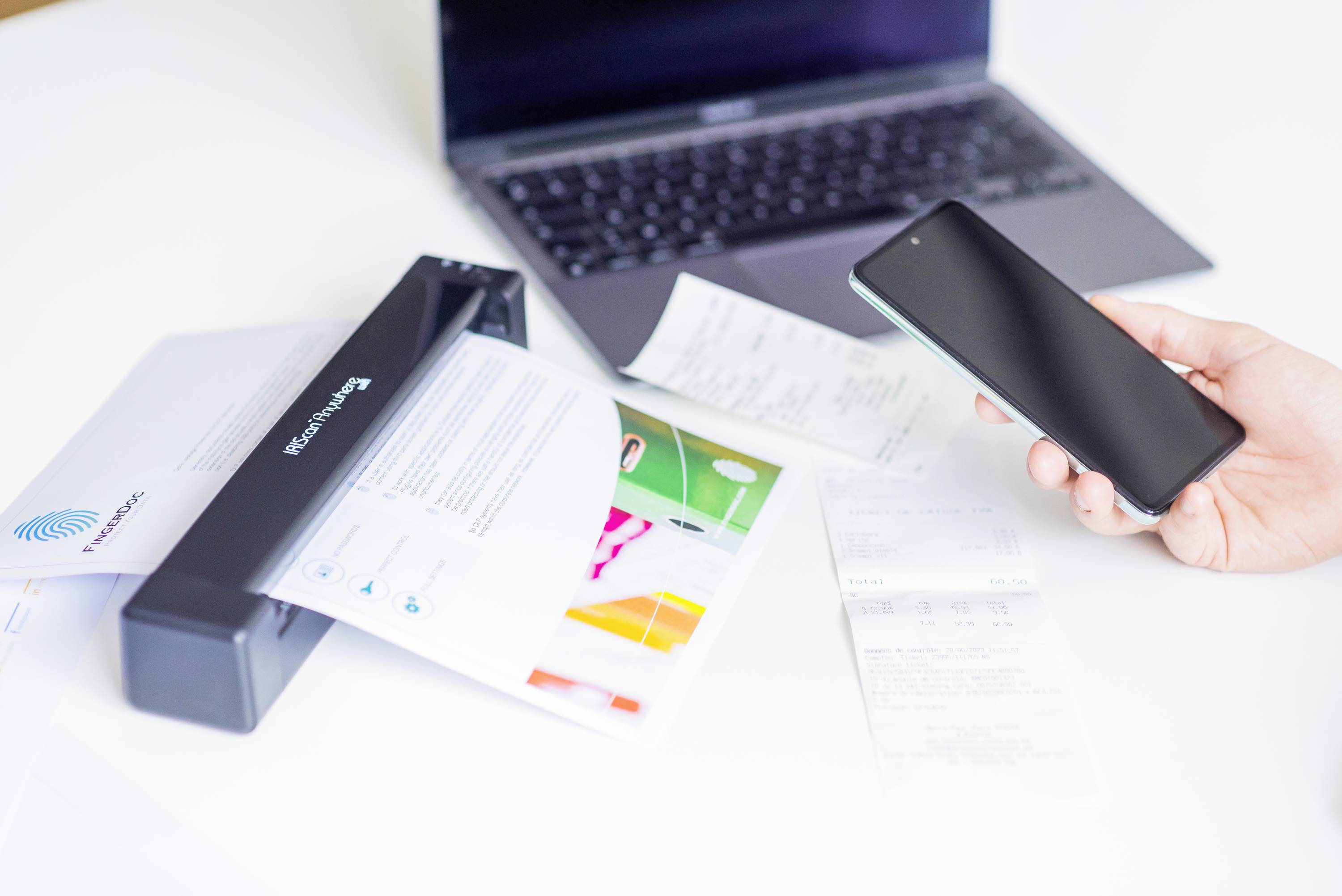 Un scanner sur une table numérise un document ; à côté se trouvent un smartphone et d'autres papiers. Un ordinateur portable est visible en arrière-plan.