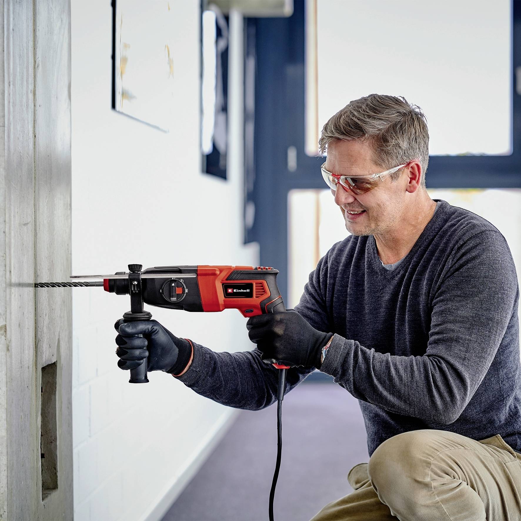 Un homme fore dans le mur avec une perceuse rouge et noire. Il porte des lunettes de protection et se réjouit de son travail.