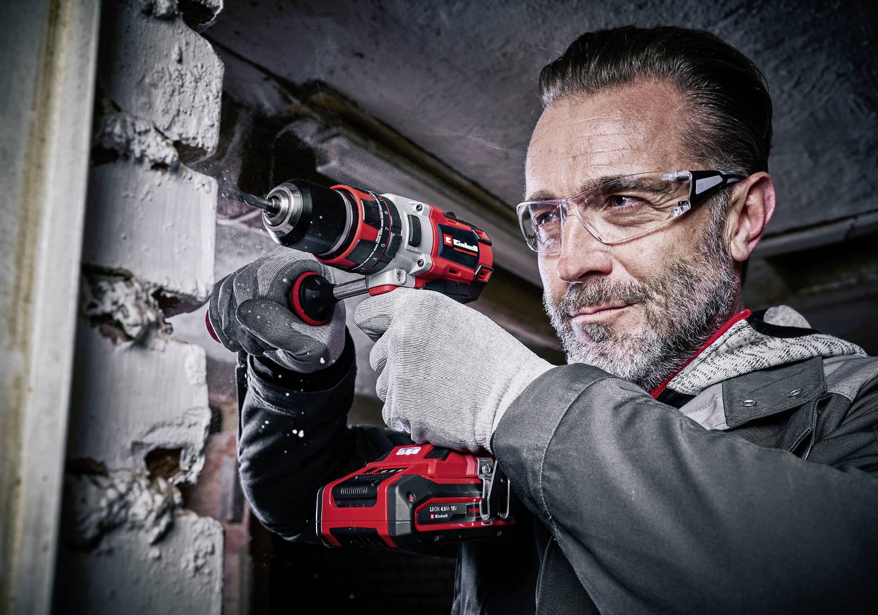 Un homme porte des lunettes de sécurité et des gants tout en utilisant une perceuse électrique rouge et noire pour travailler sur un mur de briques blanches.