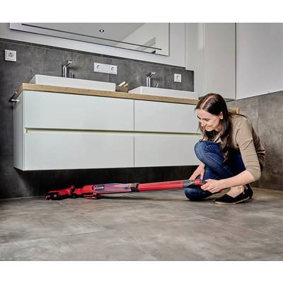 Une femme est agenouillée sur le sol de la salle de bain et nettoie la zone sous un double lavabo à l'aide d'un aspirateur sans fil rouge.