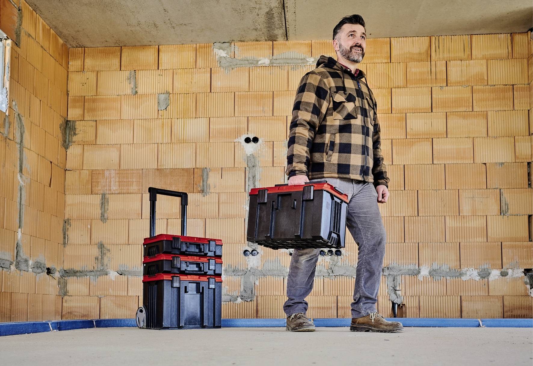 Un homme en tenue de travail porte une boîte à outils dans une pièce en construction. À côté de lui se trouve un chariot avec d'autres caisses.
