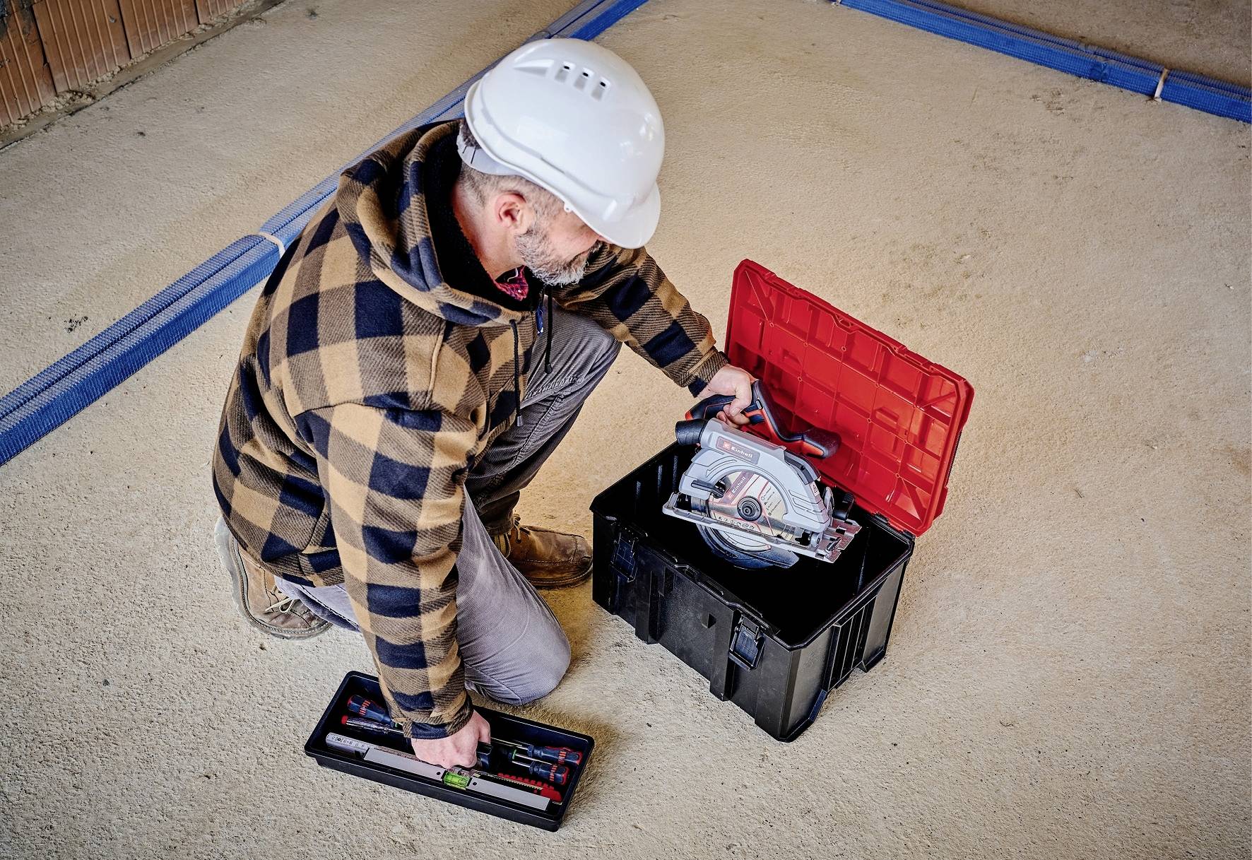 Une personne portant un casque de protection s'agenouille sur le sol, ouvre une boîte à outils et en sort une scie circulaire.