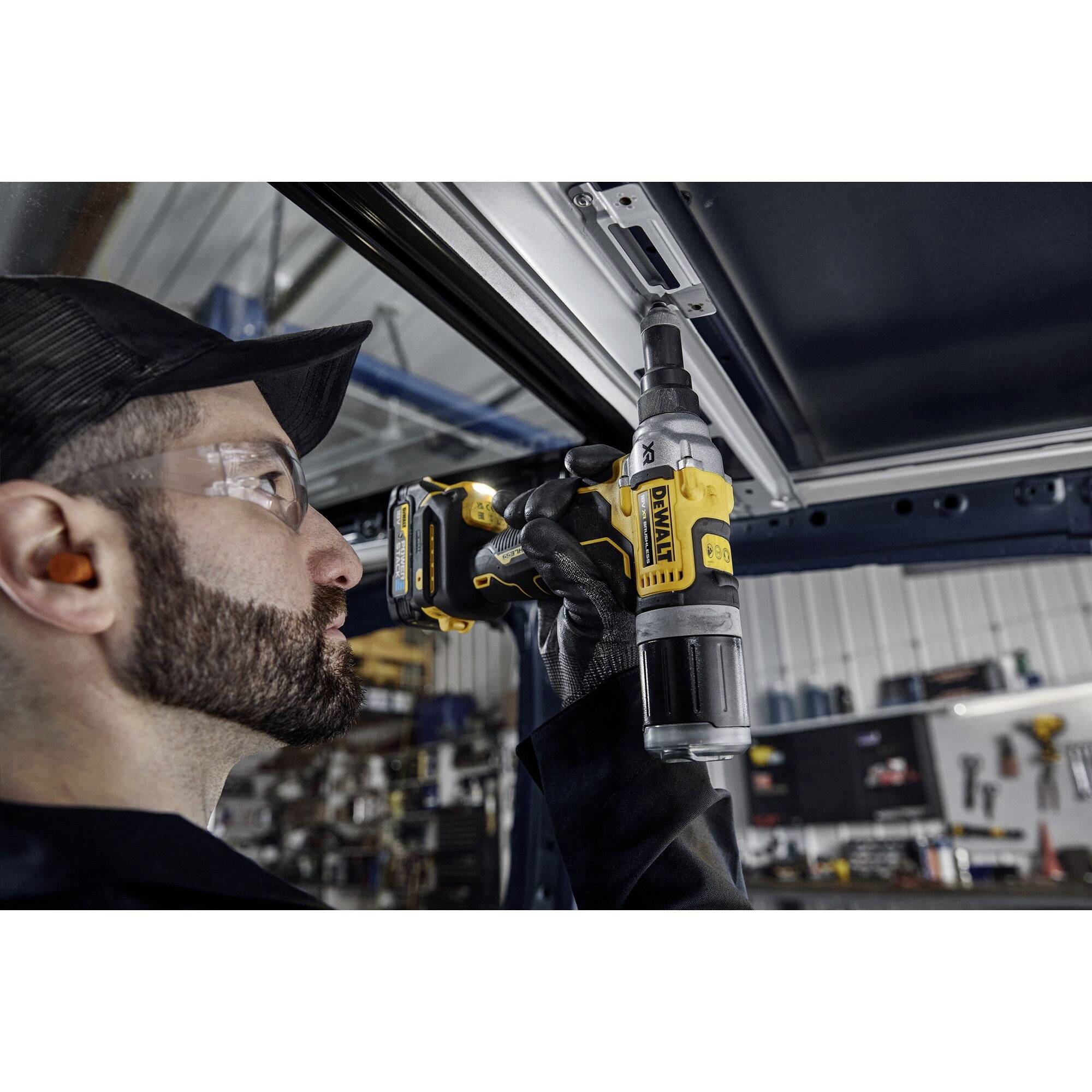 Un ouvrier portant des lunettes de protection et des protections auditives utilise une perceuse électrique pour fixer une barre métallique au plafond.