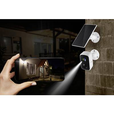 Caméra de surveillance avec panneau solaire projetant de la lumière sur un mur. Une main tient un smartphone affichant une image de surveillance d'une maison.