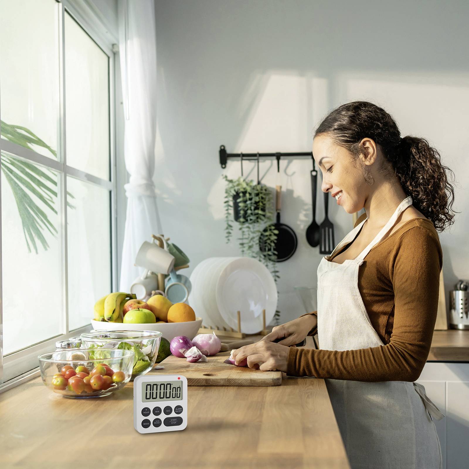 Une femme coupe des oignons dans une cuisine moderne. Sur la table, il y a un bol de tomates, un plateau de fruits agrumes et un réveil de cuisine.