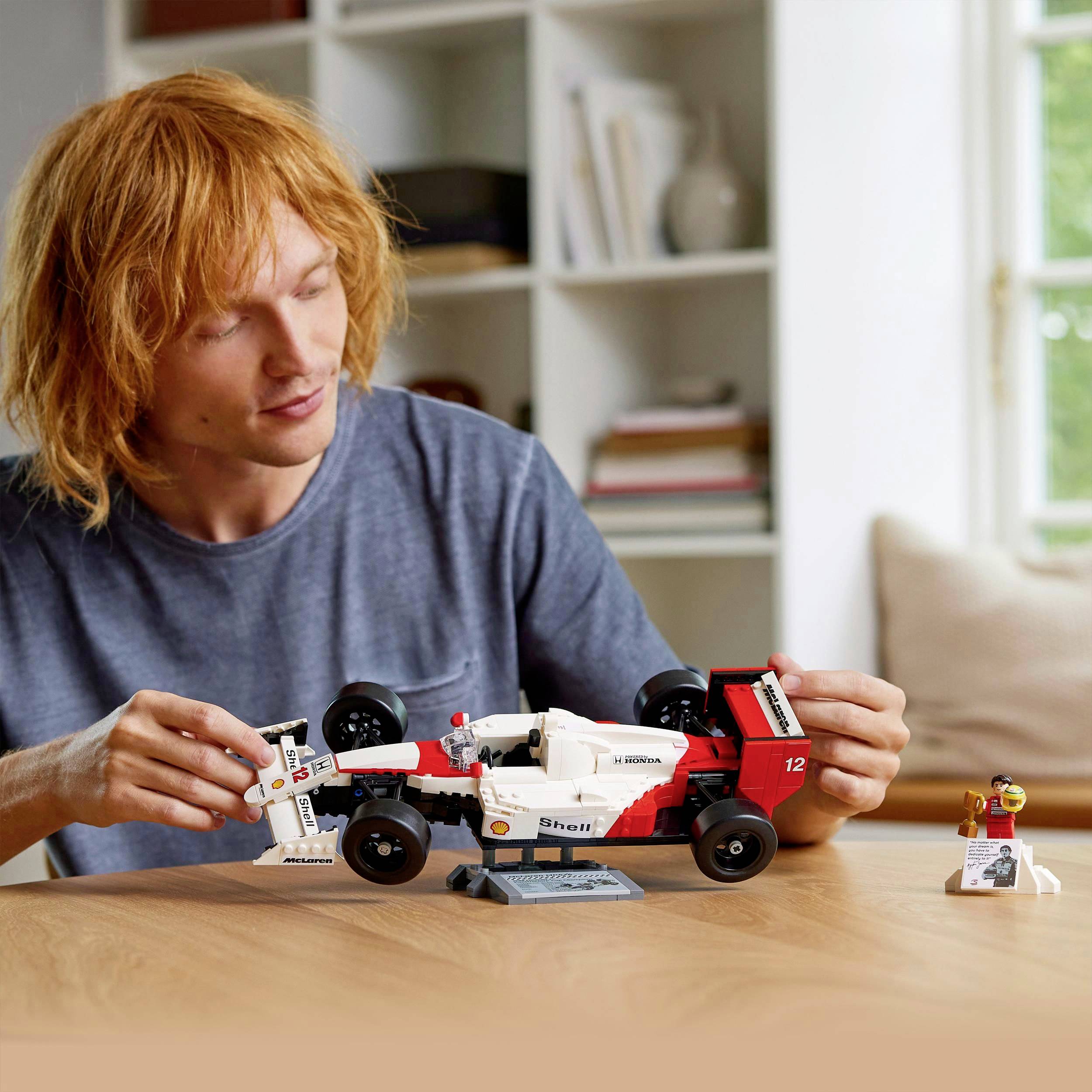 Une personne aux cheveux rouges est assise à une table et joue avec un modèle LEGO d'une voiture de course. Des étagères à livres sont visibles en arrière-plan.