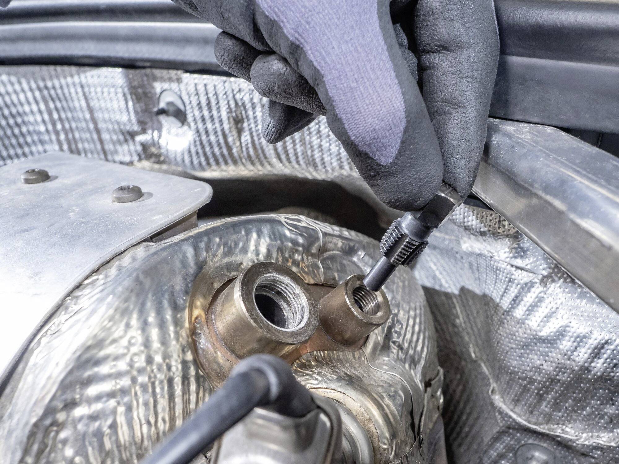 Une personne portant des gants répare ou vérifie un raccordement sur une pièce métallique sous le capot d'une voiture.