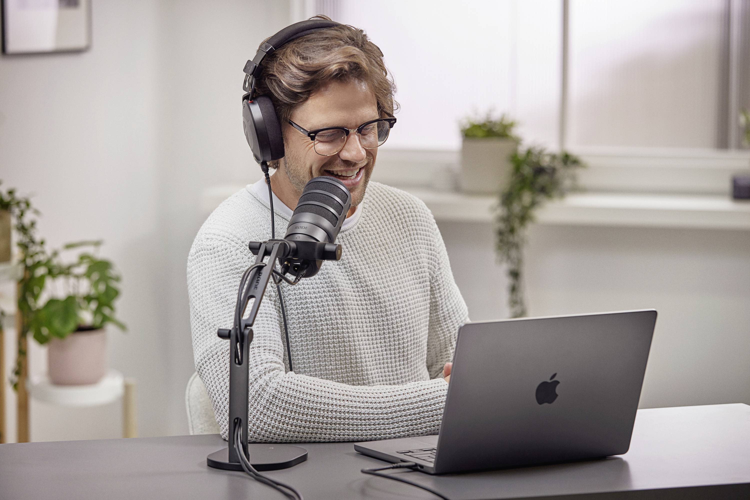 Un homme portant des lunettes et des écouteurs est assis souriant devant un microphone et un ordinateur portable dans une pièce moderne et bien éclairée.