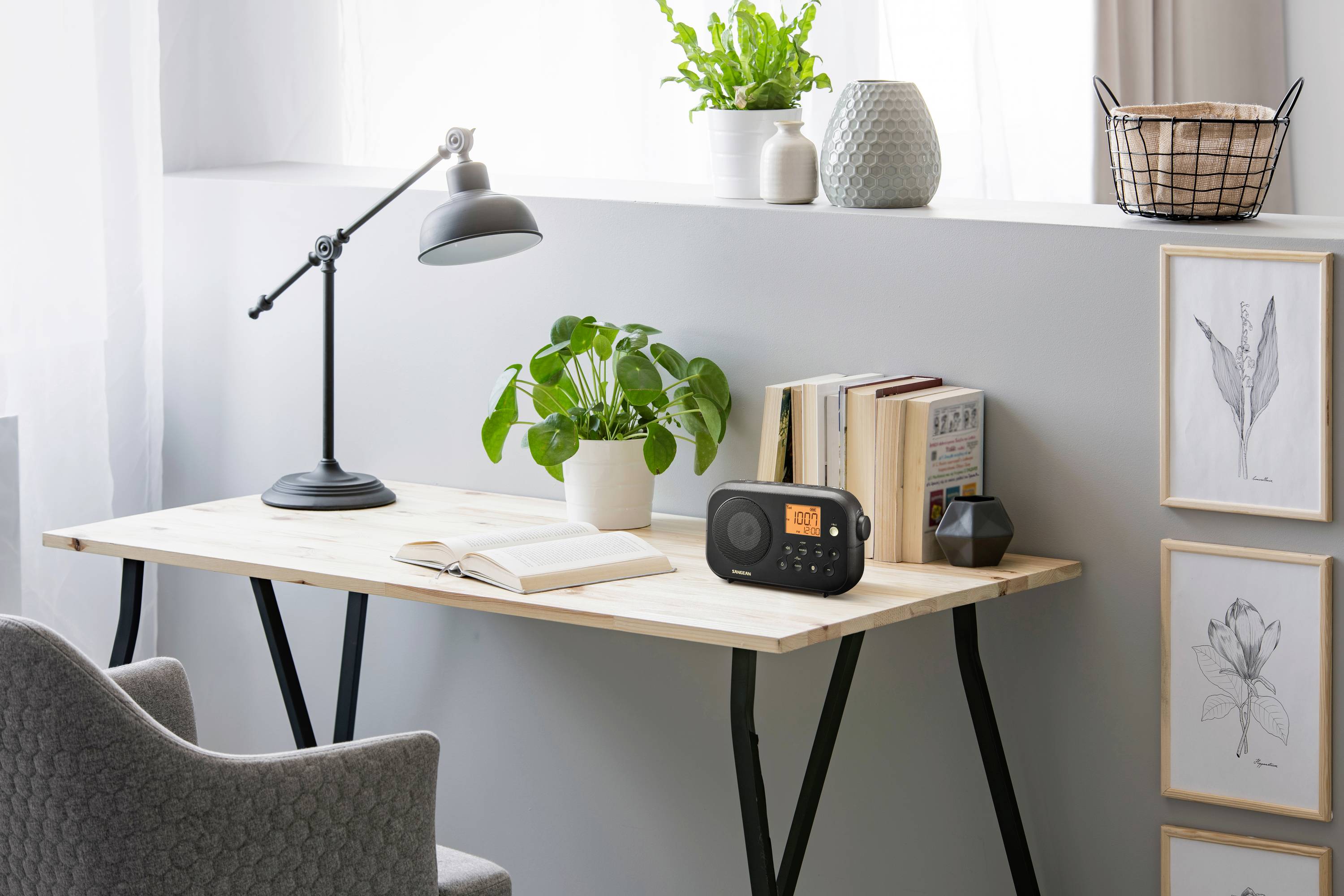 Un bureau minimaliste avec une lampe, des plantes, des livres et une radio. À côté se trouve une chaise grise.