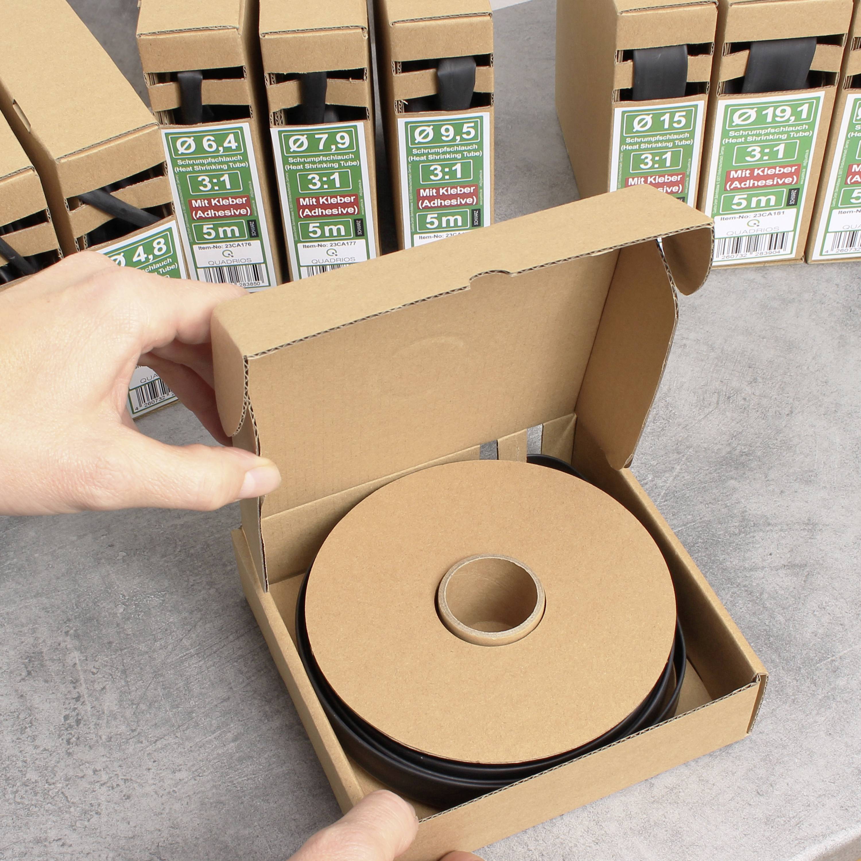 Une personne tient une boîte en carton ouverte contenant un rouleau noir en plastique. À l'arrière-plan, plusieurs boîtes similaires fermées sont disposées.