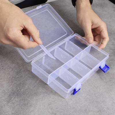 Une personne ouvre une boîte en plastique transparent vide à plusieurs compartiments sur une table grise.