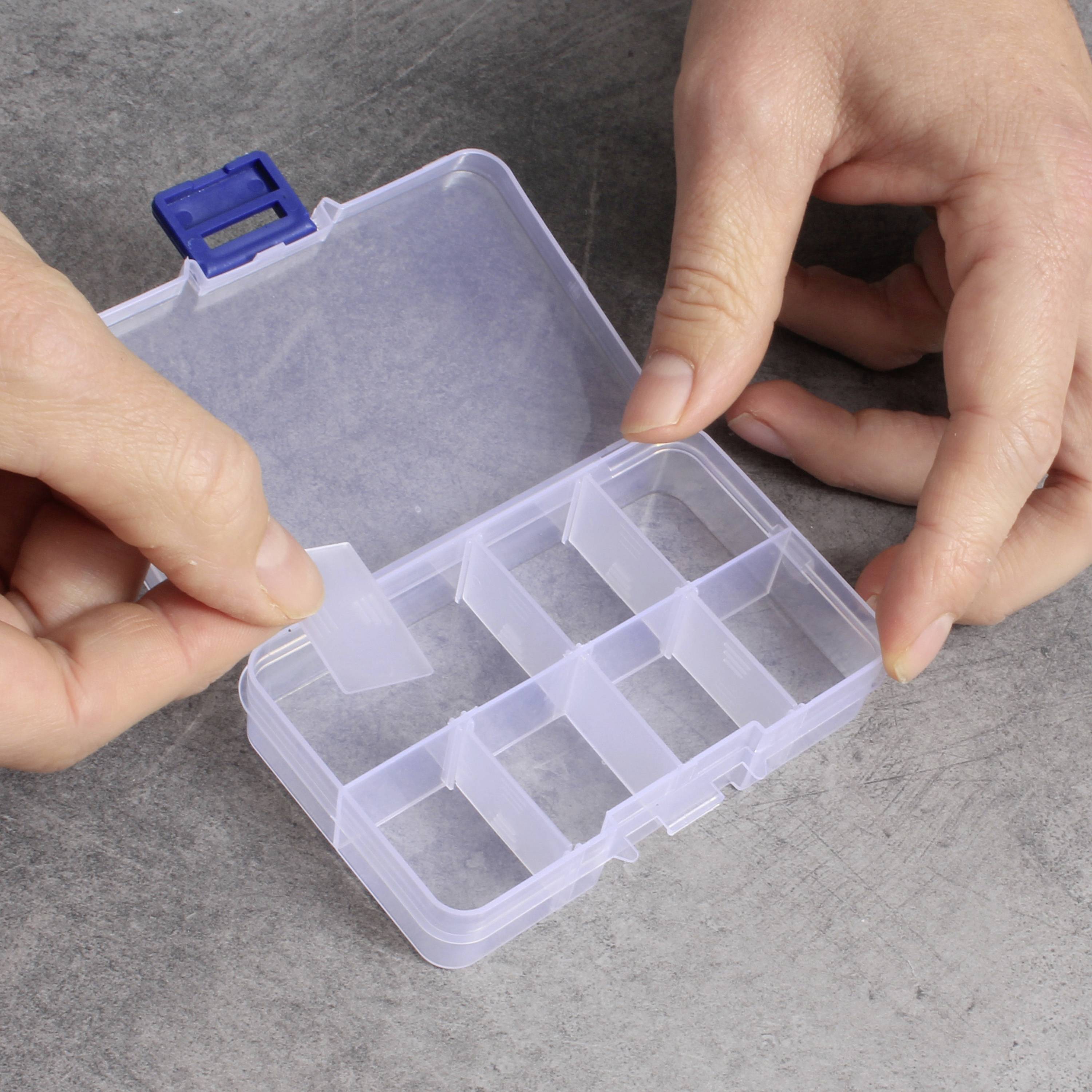 Deux mains ouvrent une petite boîte de rangement transparente à plusieurs compartiments sur une surface grise.
