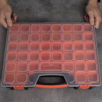 Une boîte à outils grise ouverte avec un couvercle transparent et plusieurs compartiments vides. Deux mains tiennent la boîte par ses côtés.