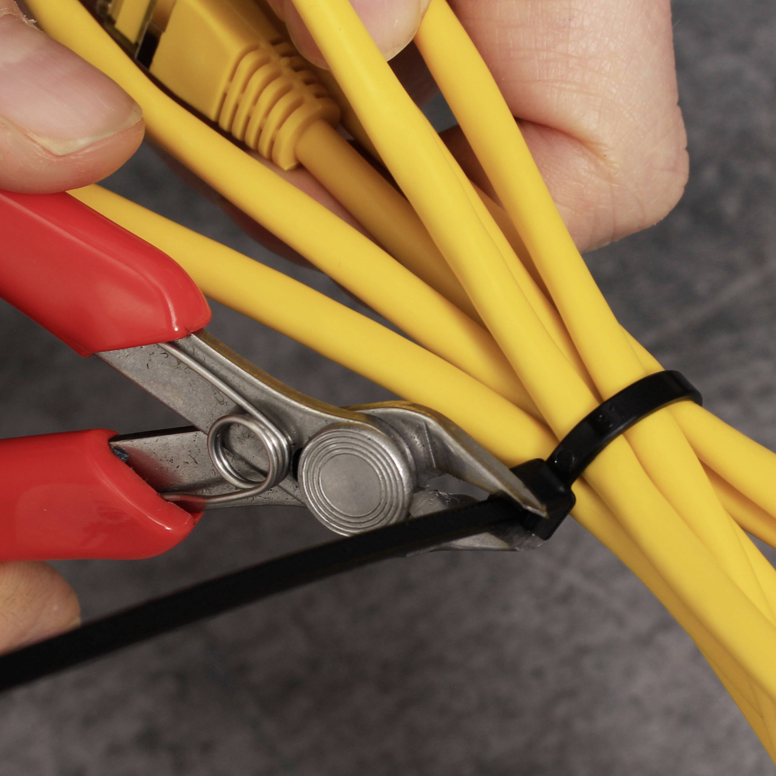 Une main tient une pince et coupe un collier de serrage noir qui maintient des câbles jaunes.