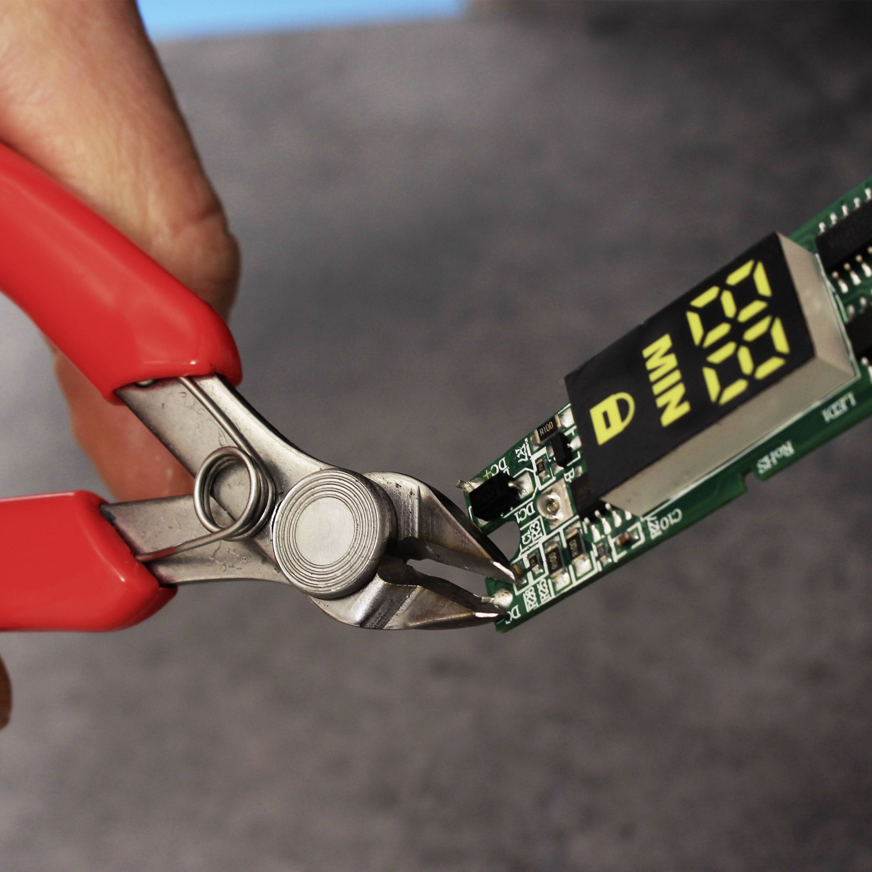 Des pinces rouges coupent une carte de circuit imprimé verte avec un affichage montrant le chiffre '21'. Les mains tiennent les pinces au-dessus d'une surface grise.