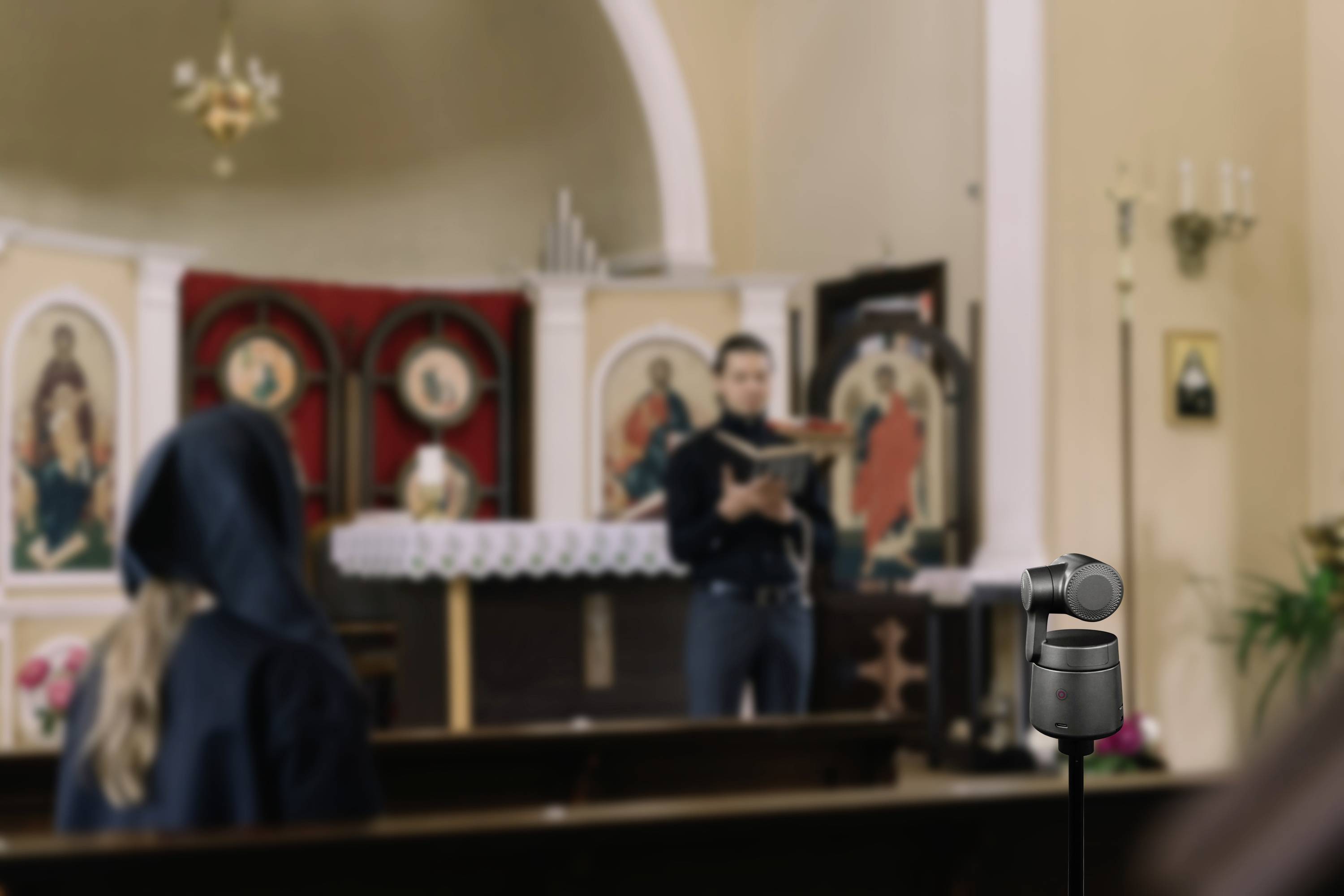 Dans une église, une personne lit un livre devant un petit groupe de personnes. Une caméra est placée au premier plan.