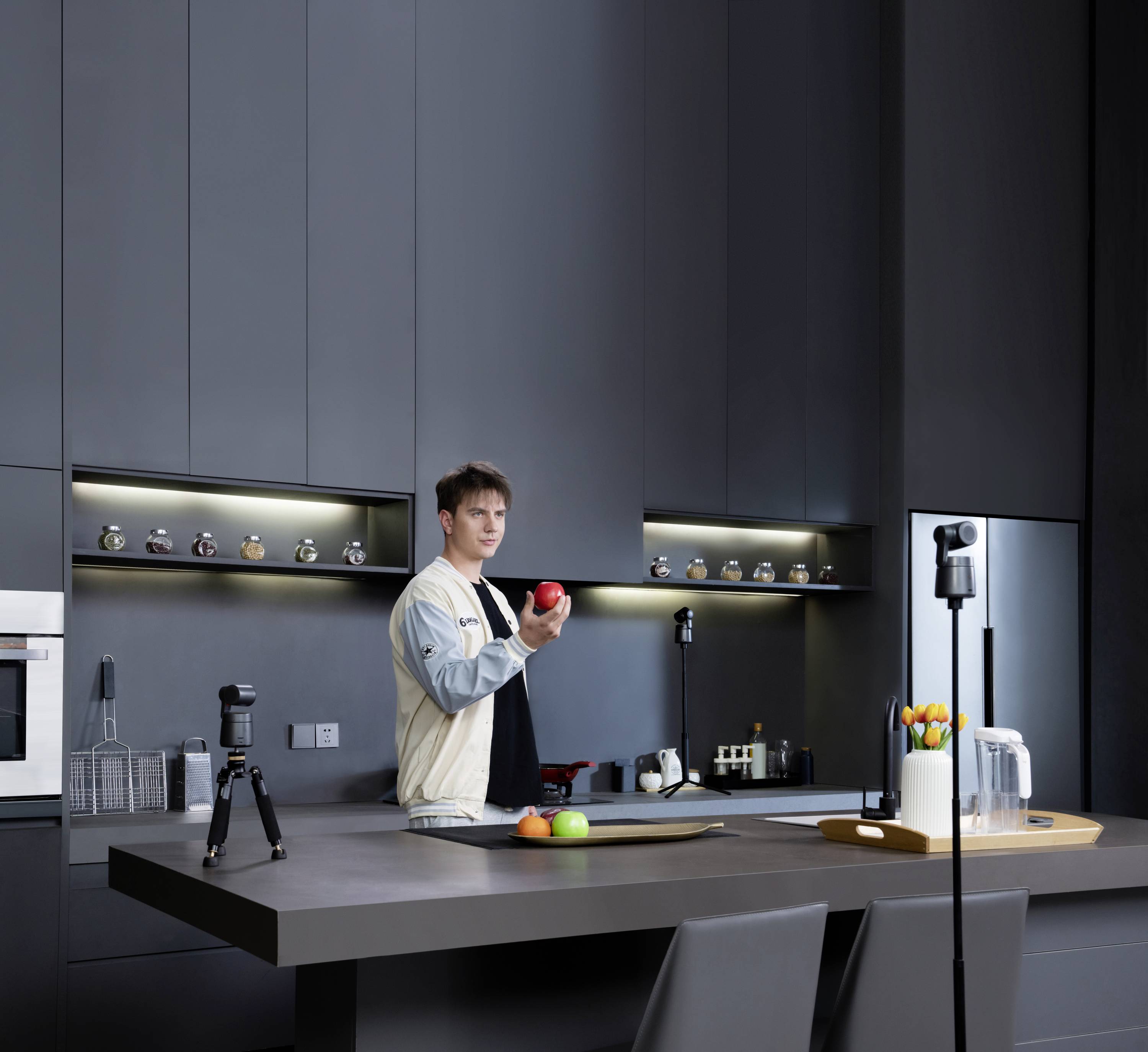 Une personne tient une pomme rouge dans une cuisine moderne au design minimaliste. Entourée de caméras et de lumières de studio.