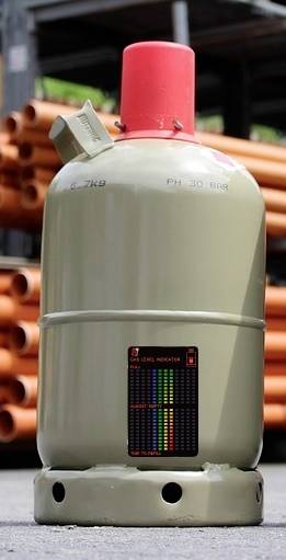 Un cylindre de gaz gris avec une valve rouge est placé devant des tubes oranges empilés. Un autocollant affiche l'échelle et les informations de sécurité.