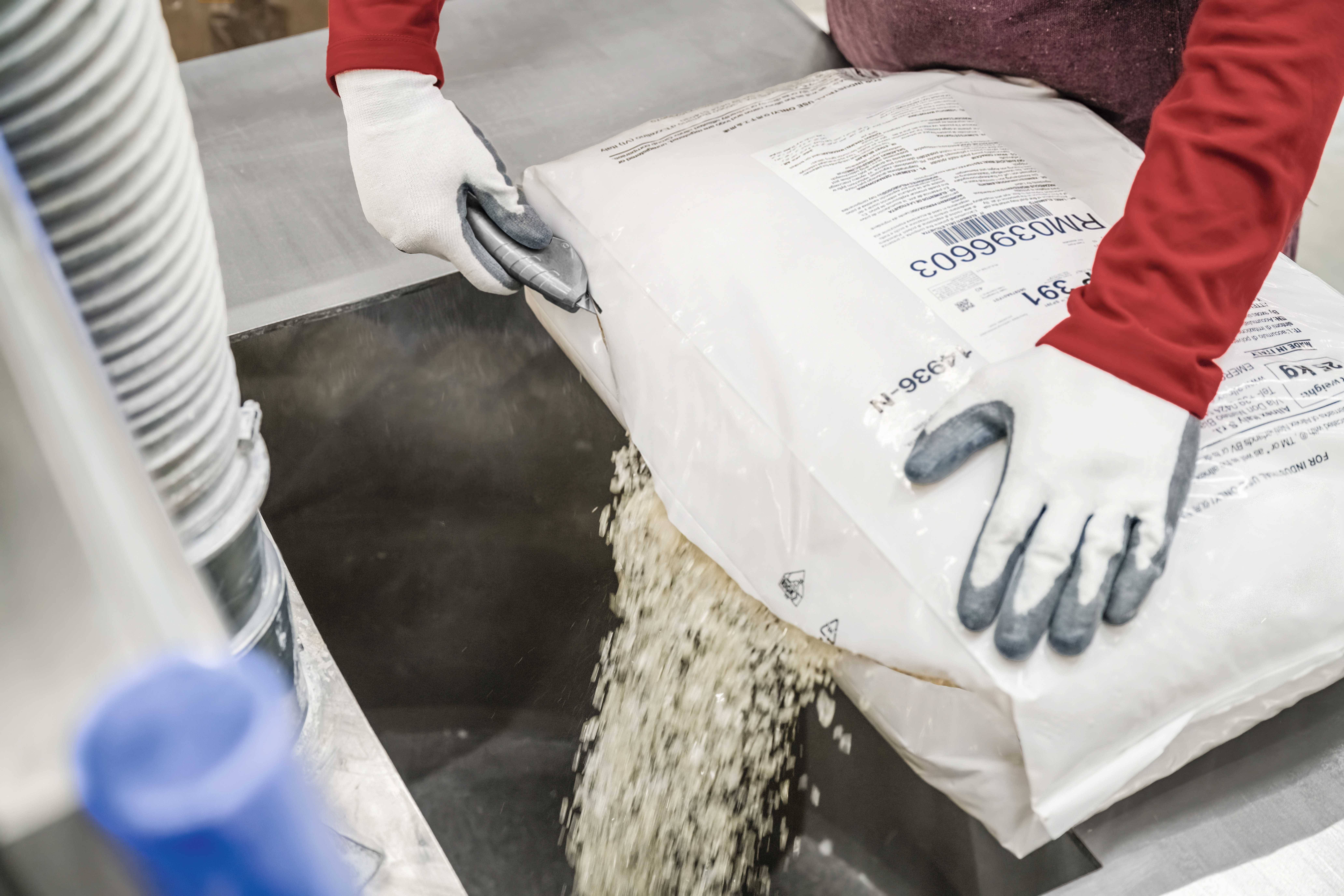 Une personne portant une chemise rouge et des gants blancs verse du riz d'un grand sac dans un conduit métallique dans un environnement industriel.