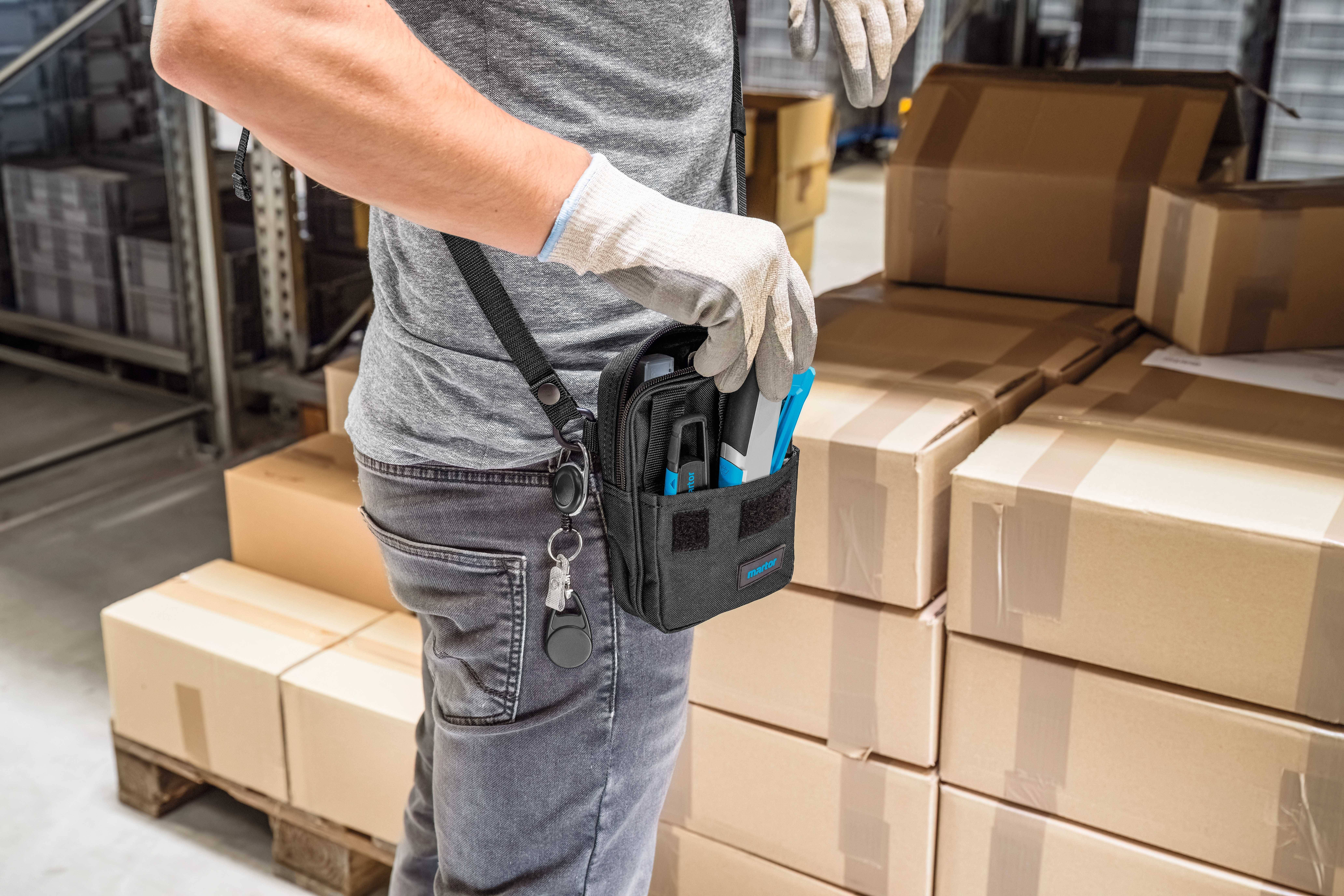 Une personne sort des outils d'un sac attaché à sa ceinture à côté de cartons empilés dans un entrepôt.