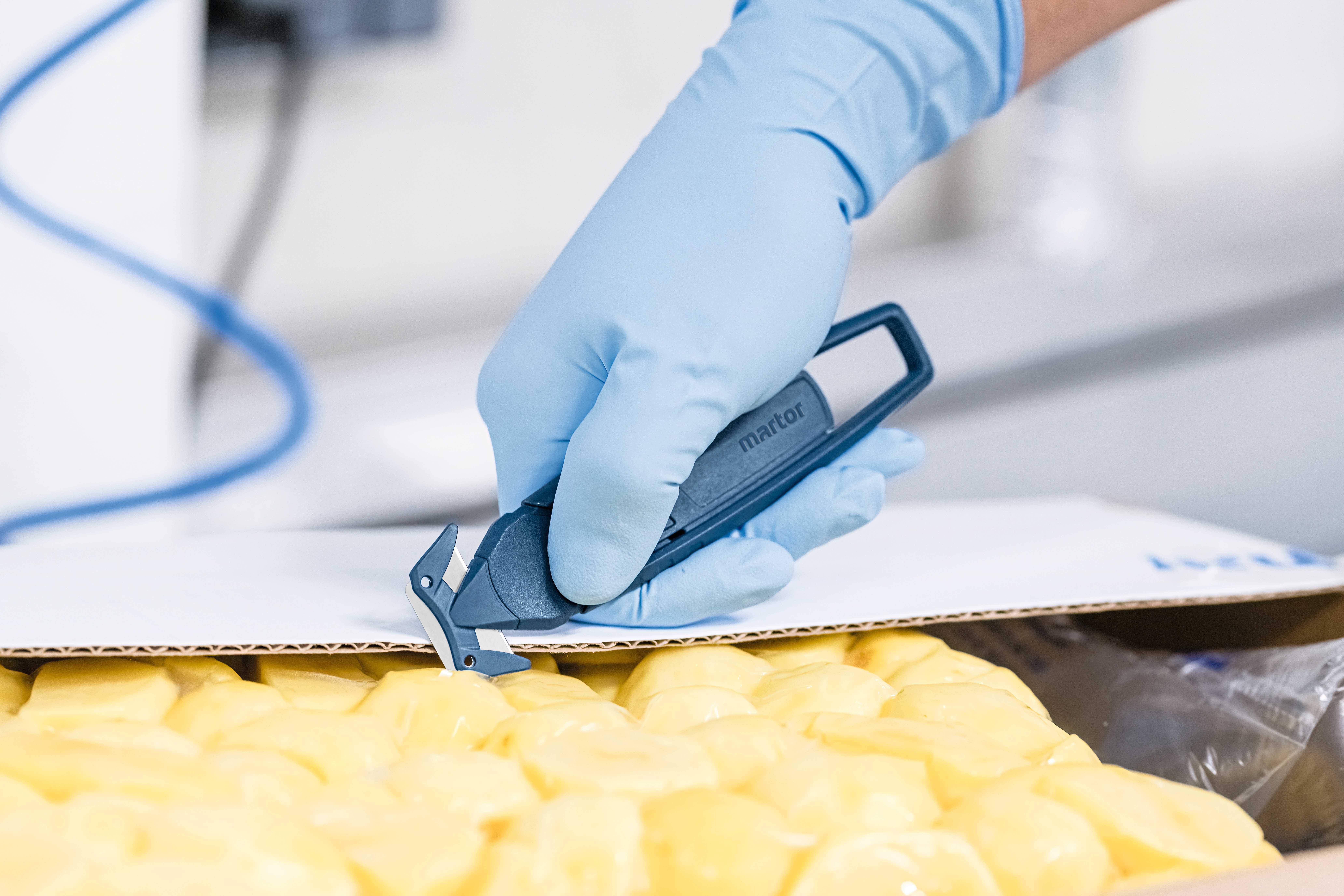 Une main gantée de bleu découpe un carton avec un couteau de sécurité, rempli d'objets emballés en jaune.