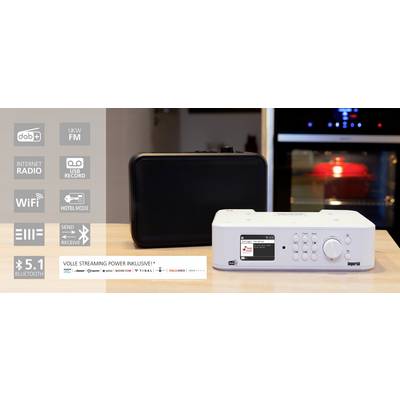 Radio numérique avec de nombreuses fonctionnalités posé sur une table de cuisine, à côté d'un boîtier noir. En arrière-plan, un four est visible.