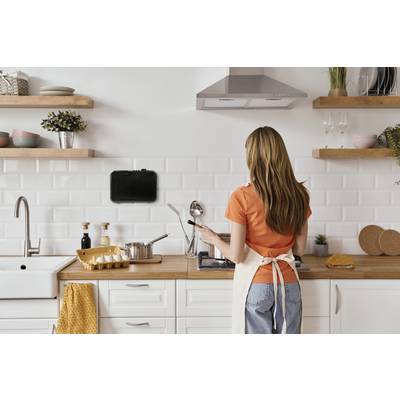 Femme portant un t-shirt orange et un tablier, cuisinant dans une cuisine moderne. Elle utilise une tablette comme guide de recette accroché au mur.