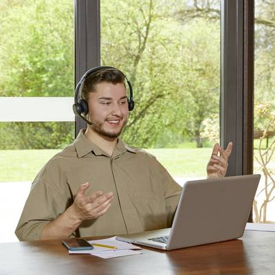 Un homme est assis souriant devant un ordinateur portable, portant des écouteurs, faisant des gestes avec sa main, entouré d'un jardin vert visible par la fenêtre.