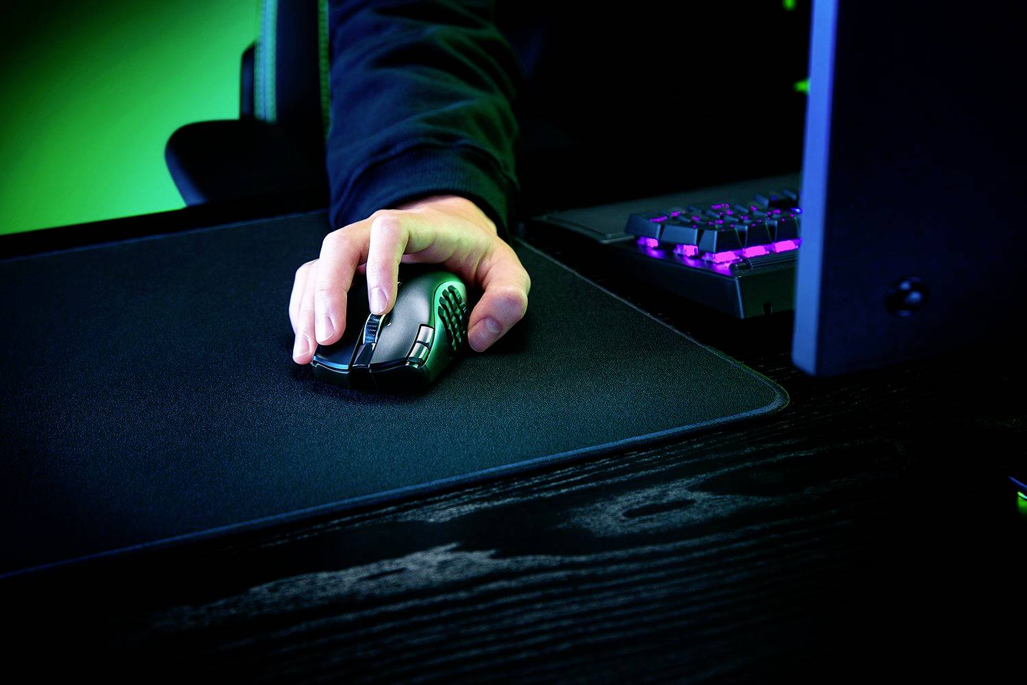 Une main manipule une souris de gaming sur un tapis de souris. En arrière-plan, on aperçoit des parties d'un clavier de gaming rétroéclairé et un écran.